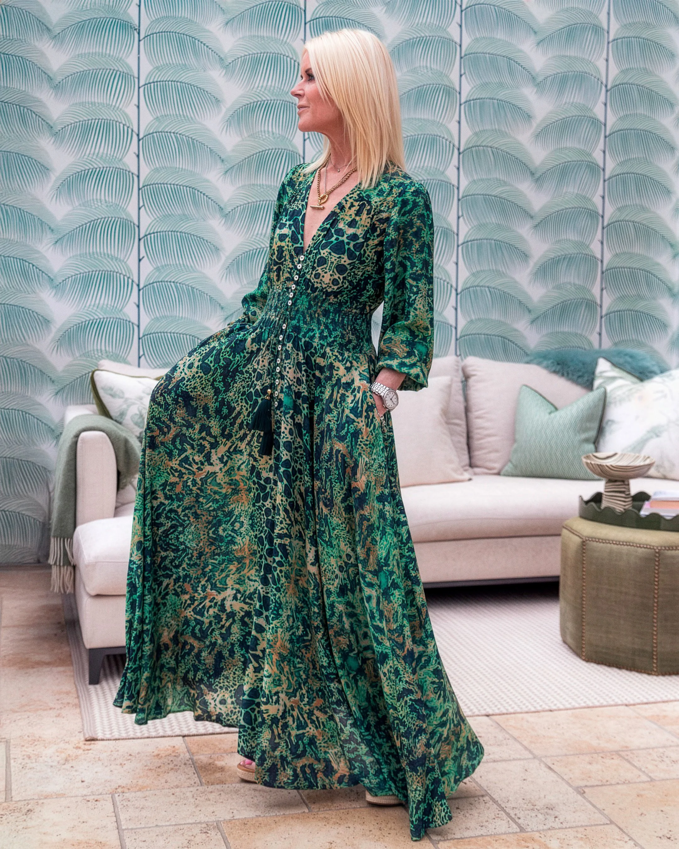 A woman with blonde hair wearing a long, green animal print dress standing indoors near a pastel-colored sofa and patterned wallpaper.