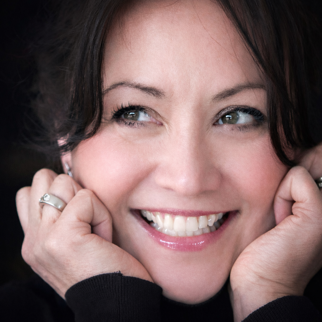 Close-up of a woman with dark brown hair, smiling with teeth showing, touching her face with both hands, wearing jewelry.