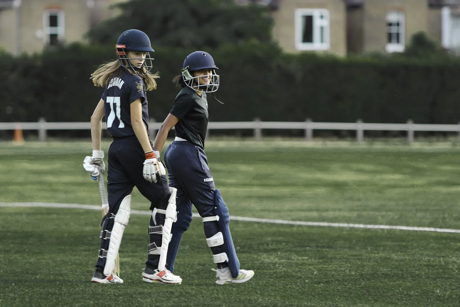 Two young female cricket players walking on a cricket field, wearing helmets, gloves, and protective gear, with a background of green grass and houses.