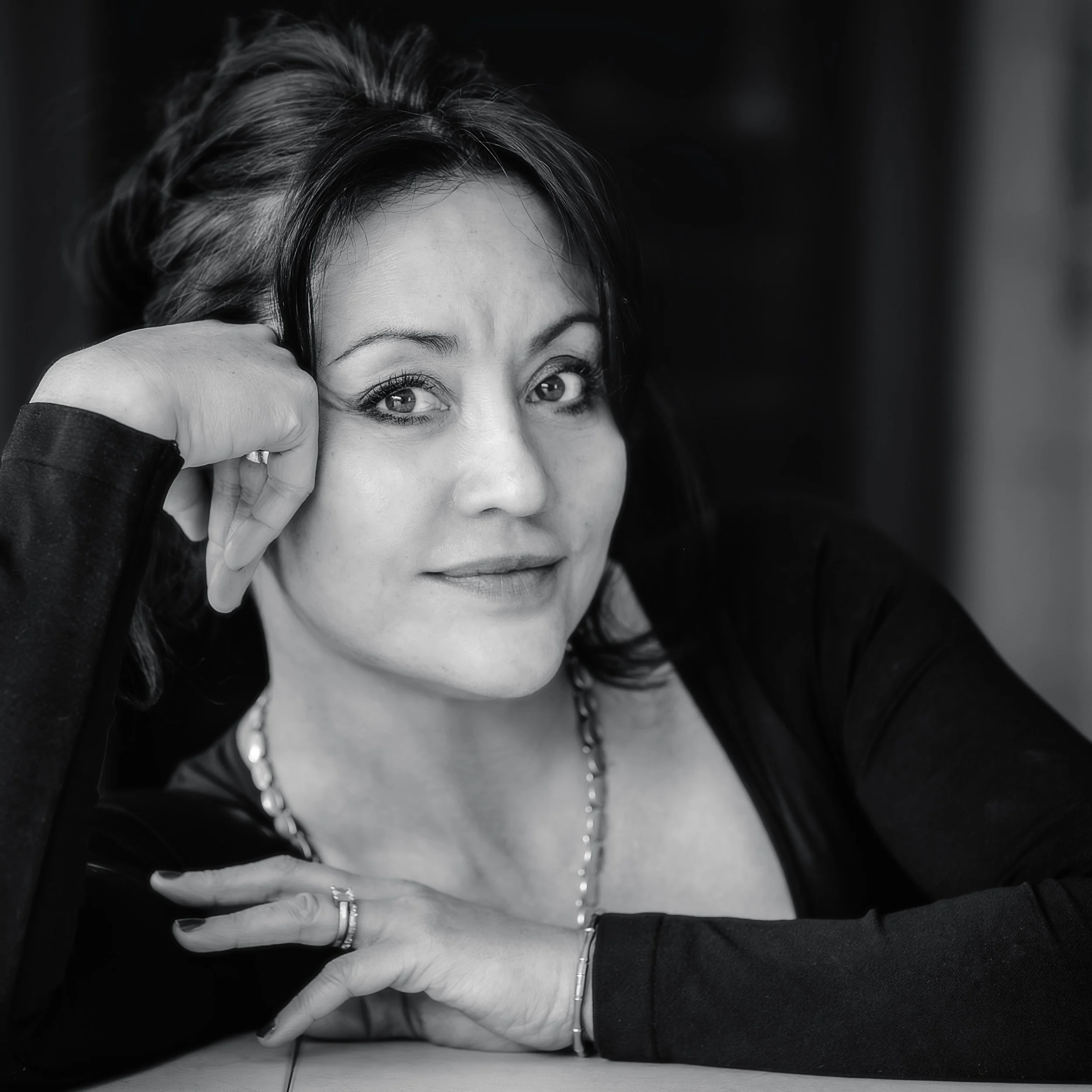 Black and white portrait of a woman with dark hair, wearing a necklace and a ring, resting her head on her hand and smiling gently.