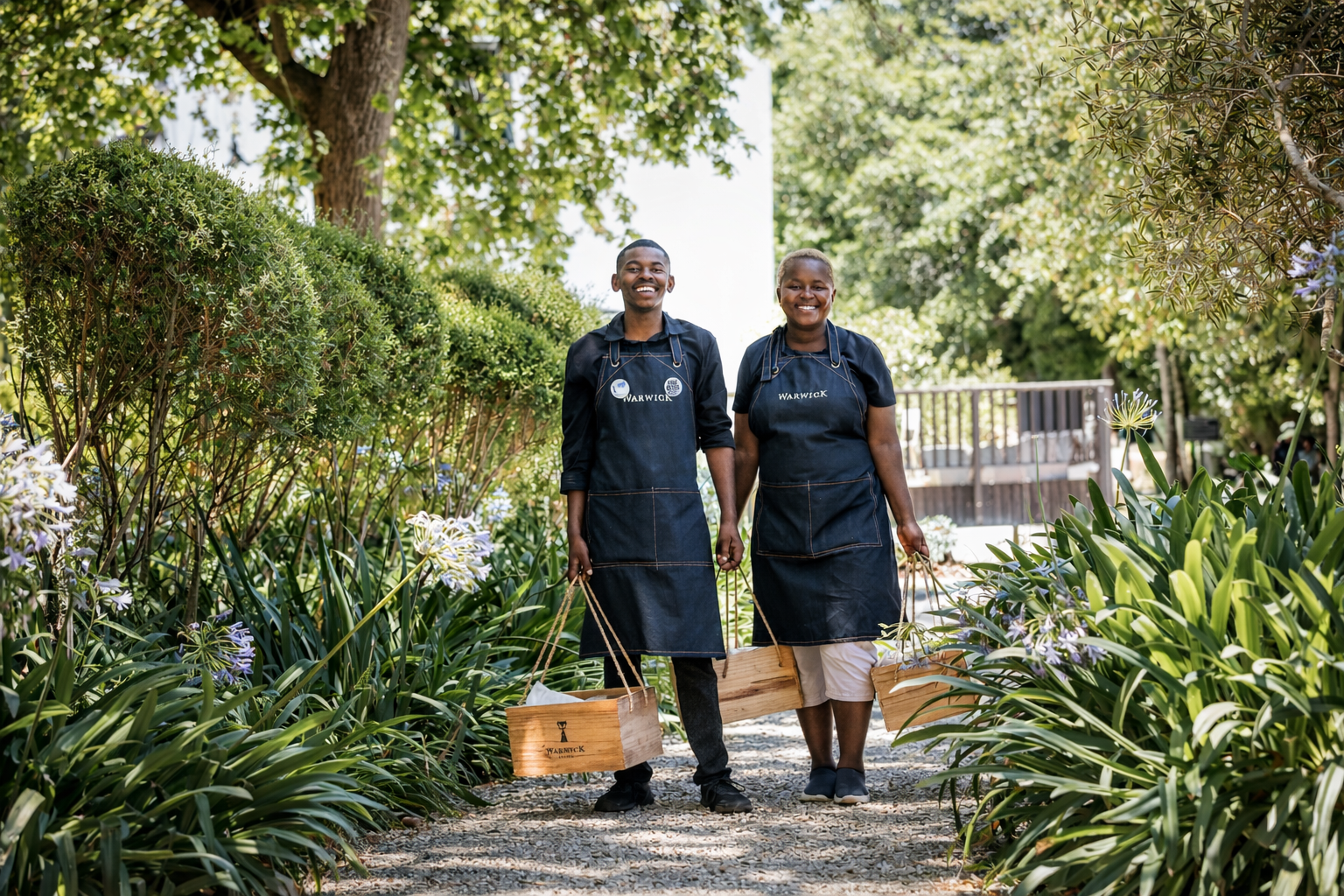 Two smiling people in black aprons walking through a garden with green plants and trees, holding bags.