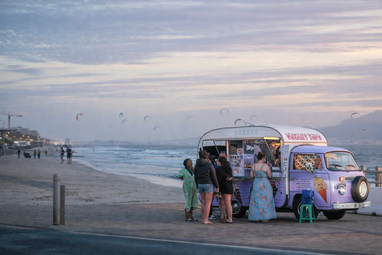 A colorful ice cream truck parked on a beach promenade during sunset, with several people gathered around ordering ice cream.