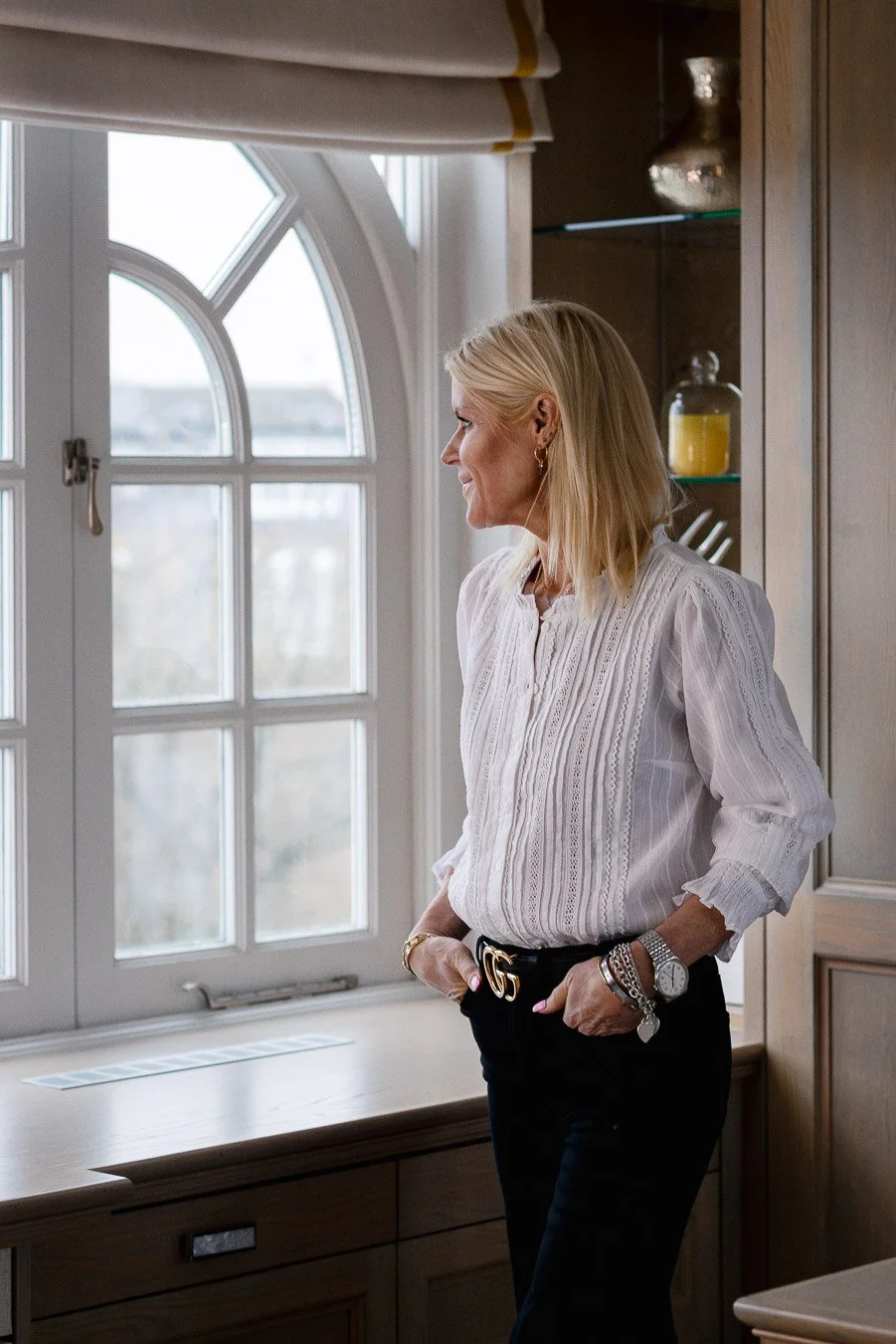 A woman with blonde hair, wearing a white blouse and black pants, standing in a room by a window, looking outside with her hands in her pockets.
