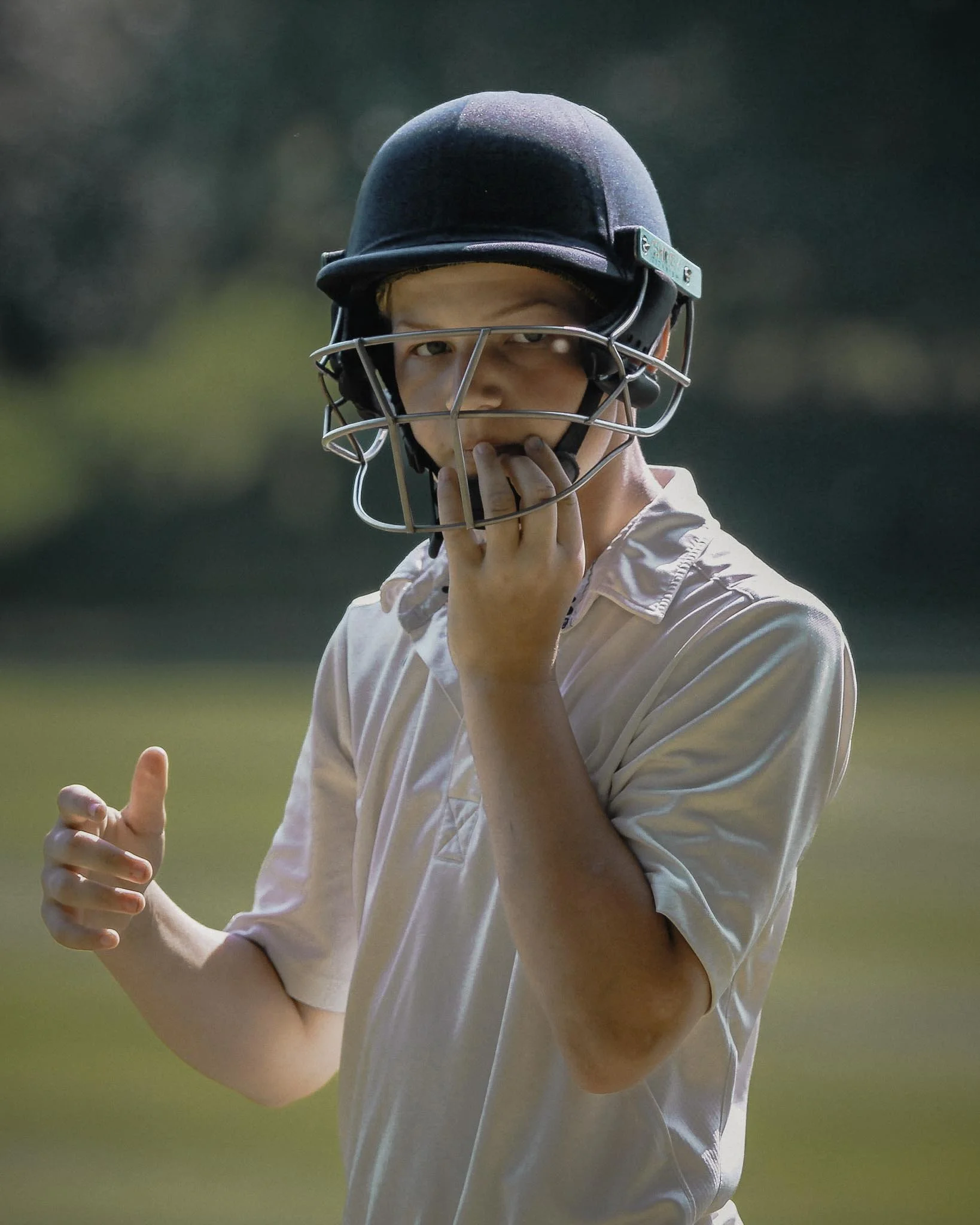 A young boy wearing a beige shirt and a black football helmet with a face guard, standing outdoors on a grassy field with trees in the background, touching his face guard with his right hand.