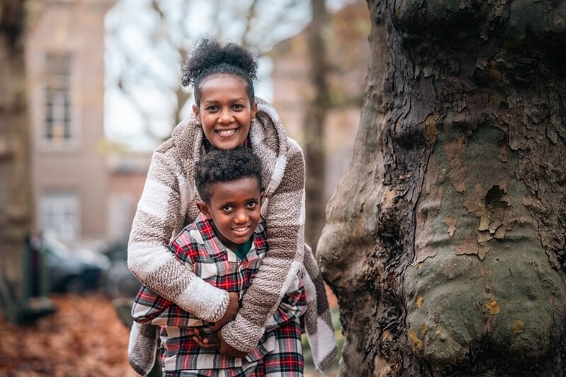 A smiling woman hugging a young boy outdoors near a large tree with autumn leaves on the ground.