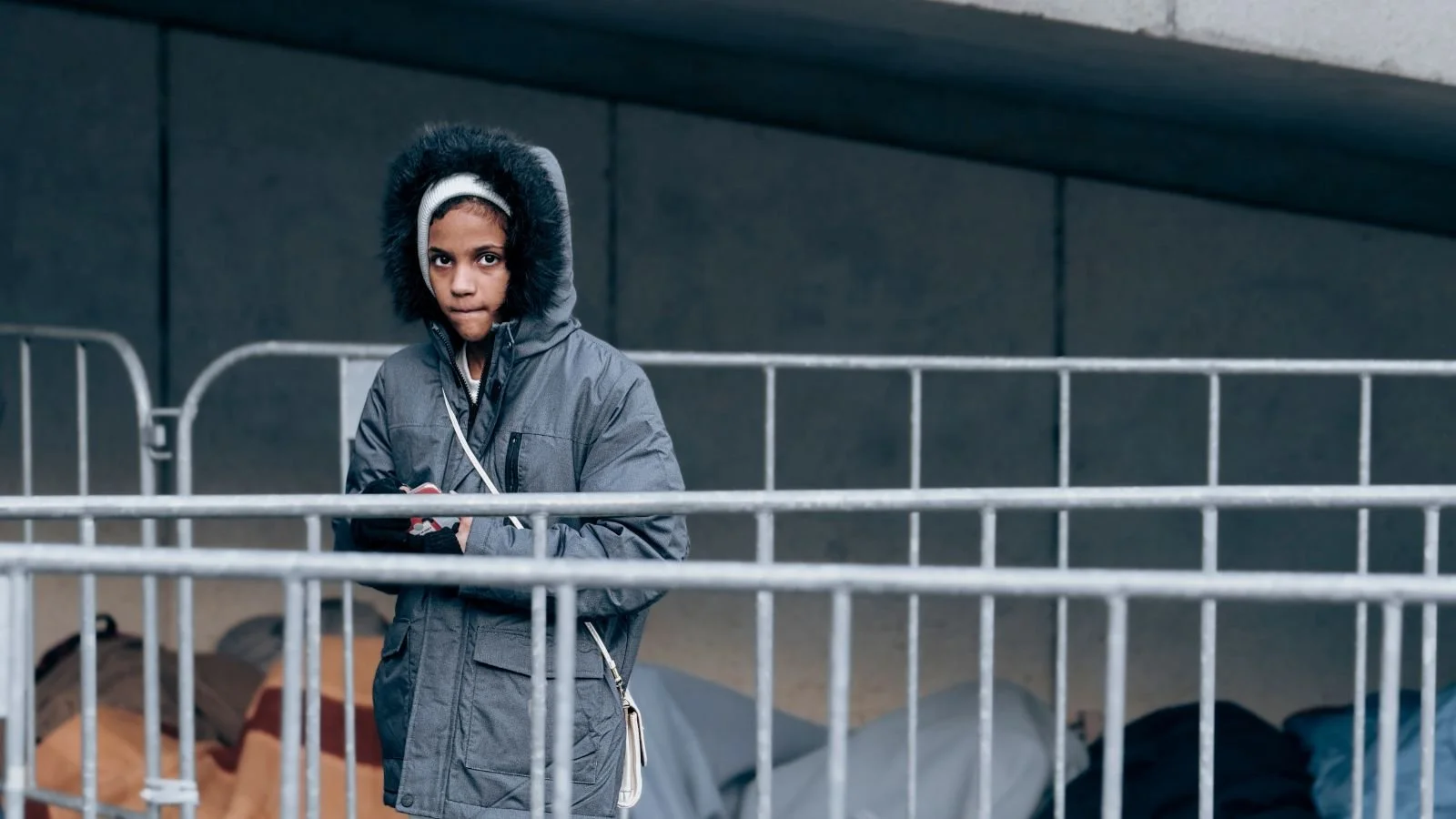 A young boy with dark curly hair wearing a gray winter coat with a fur-lined hood, standing behind a metal railing in an outdoor urban setting with tents and backpacks in the background.