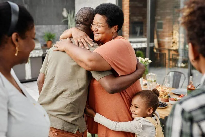 A woman and a man hug warmly, surrounded by smiling people at an outdoor gathering with a table of food in the background.