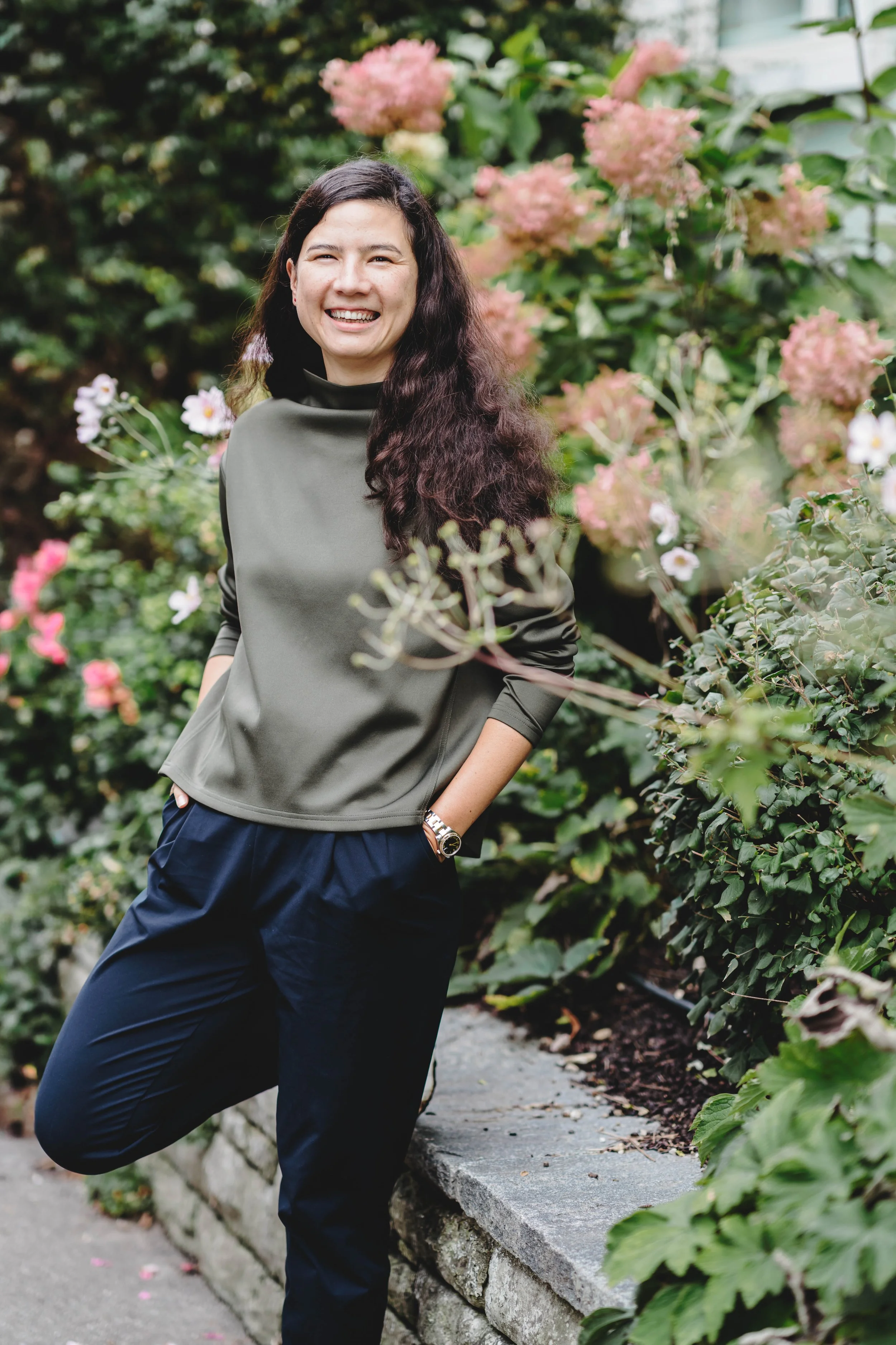 Lea Im Obersteg with long dark hair, smiling, wearing a green pullover and dark pants, standing outdoors near a stone wall with pink and white flowers in the background, welcoming you on the website.