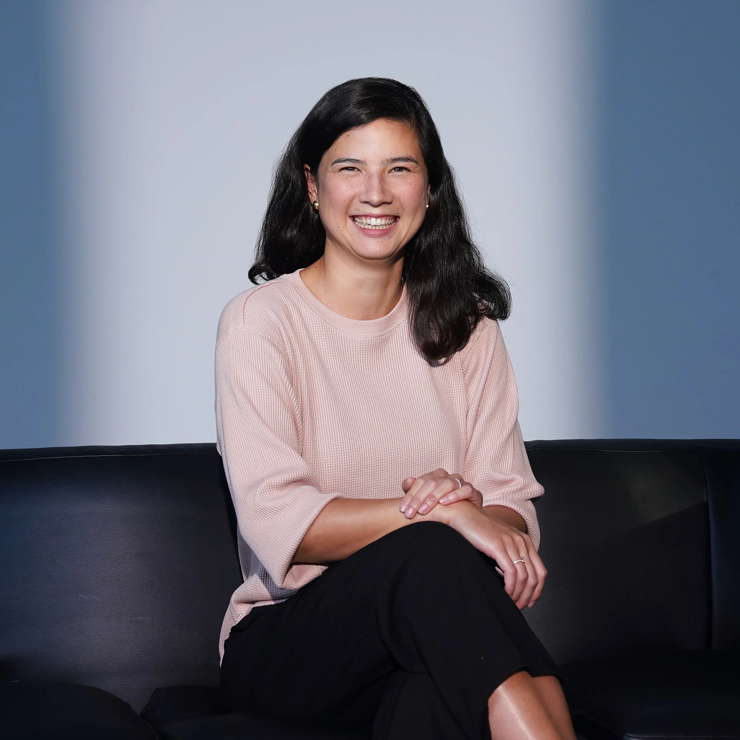 A picture of Lea Im Obersteg with dark hair, wearing a light pink top and black pants, sitting on a black leather couch with her hands clasped and smiling at the camera.