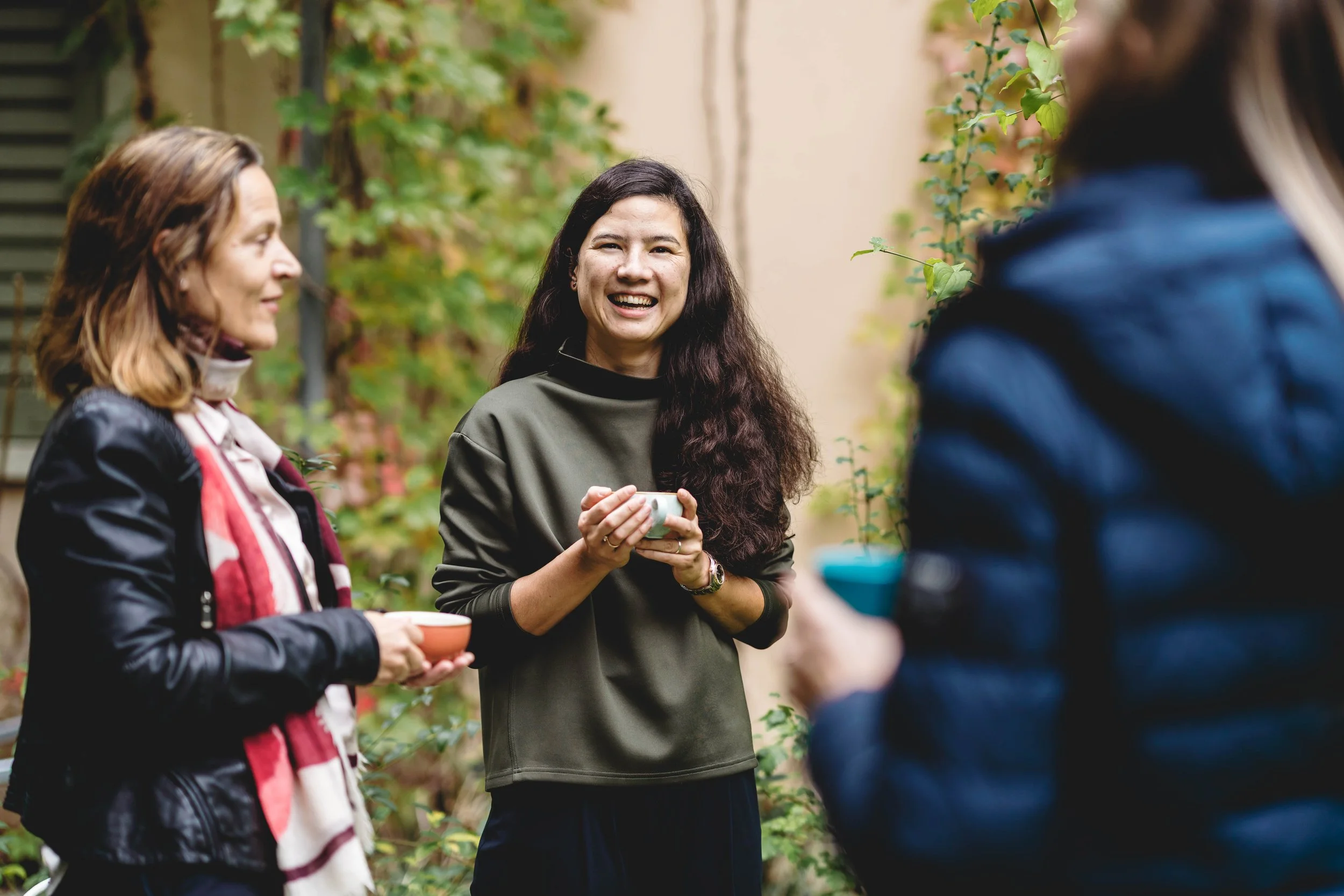 Lea Im Obersteg with long dark hair smiling and holding a cup, in a garden setting with green plants and a beige wall in the background, representing a moment to connect over a cup of coffee.