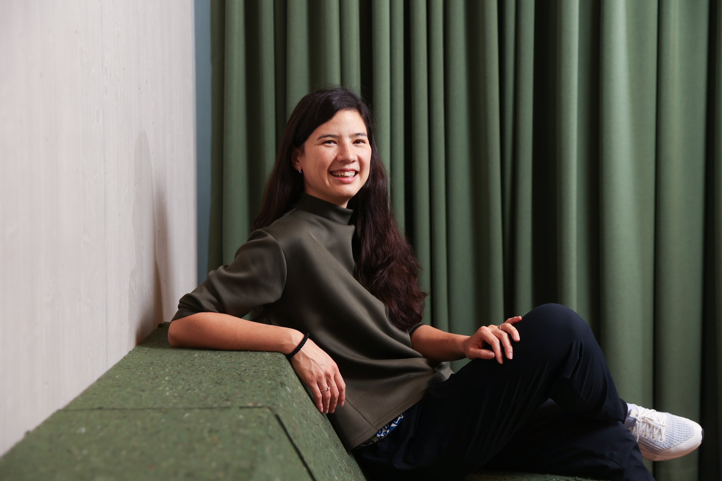 Lea Im Obersteg, wearing a dark green pullover, sitting on a green carpeted bench in front of green curtains, smiling at the camera and inviting you to connect.