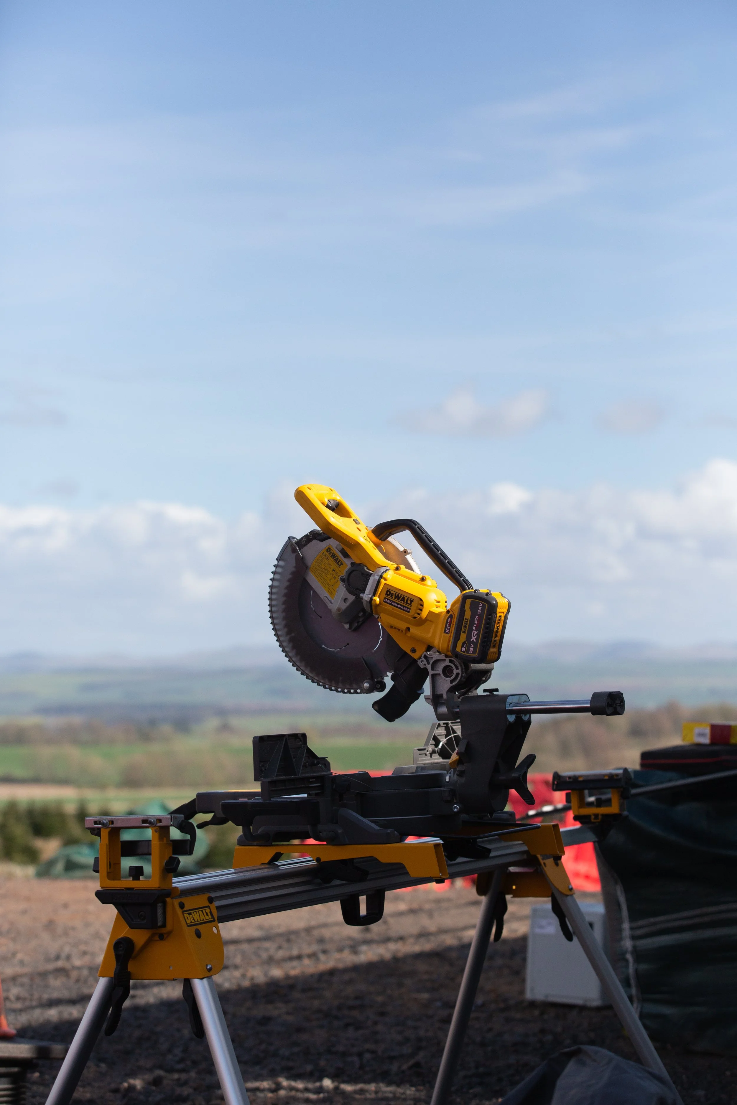 A yellow DEWALT laser saw equipped with a circular saw blade is mounted on a yellow adjustable stand outdoors. The background features a landscape with a blue sky and scattered clouds.