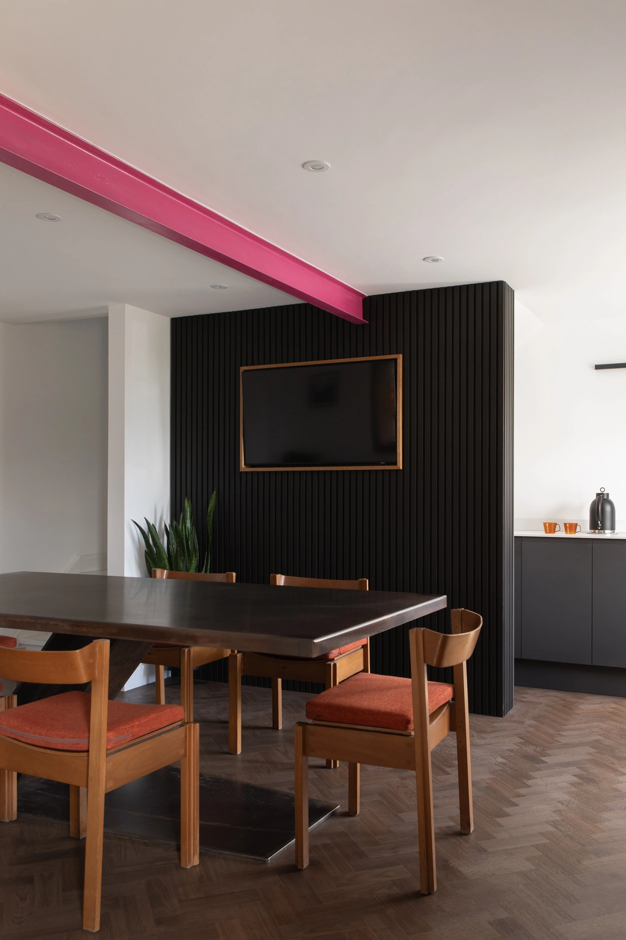 Modern dining room with a dark wooden table, four wooden chairs with orange cushions, a black textured accent wall with a mounted flat-screen TV, green plant, and a kitchen counter with two orange cups and a silver coffee pot in the background.