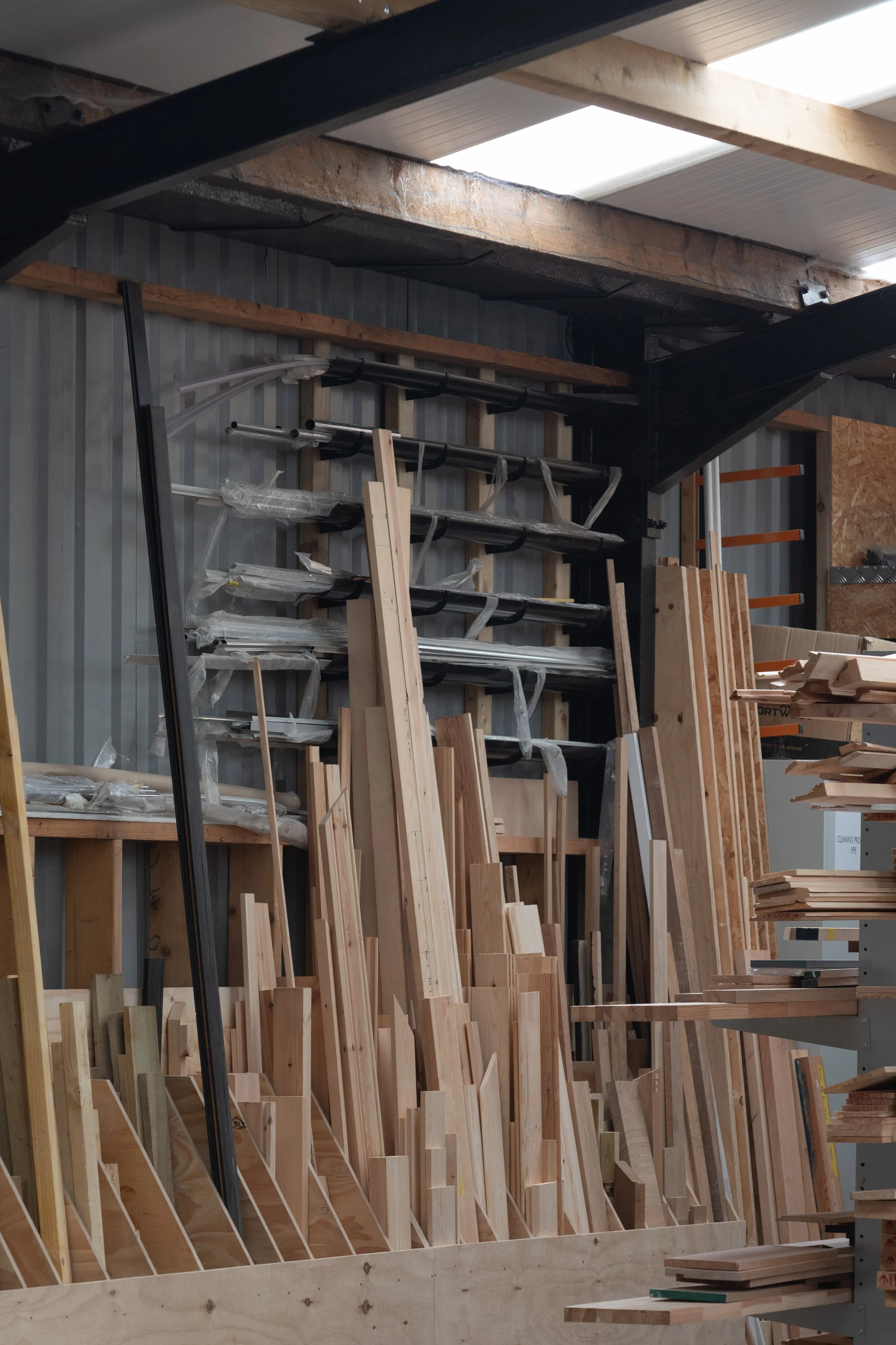 Wooden furniture pieces and planks arranged on shelves in a woodworking shop, with unfinished wood structures and tools visible.