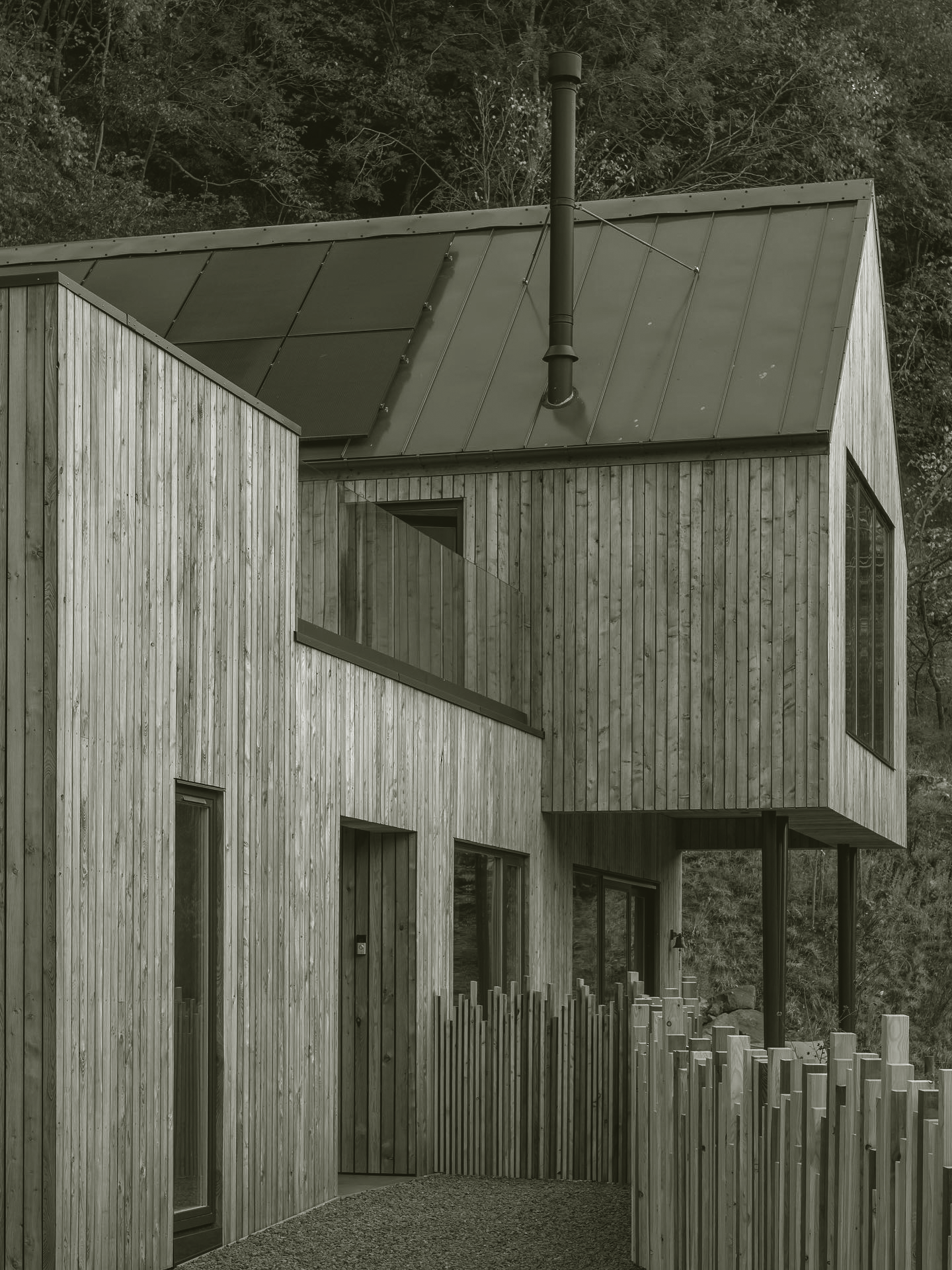A modern house with wooden siding and large windows, set against a wooded hillside, with a metal roof and a chimney pipe.