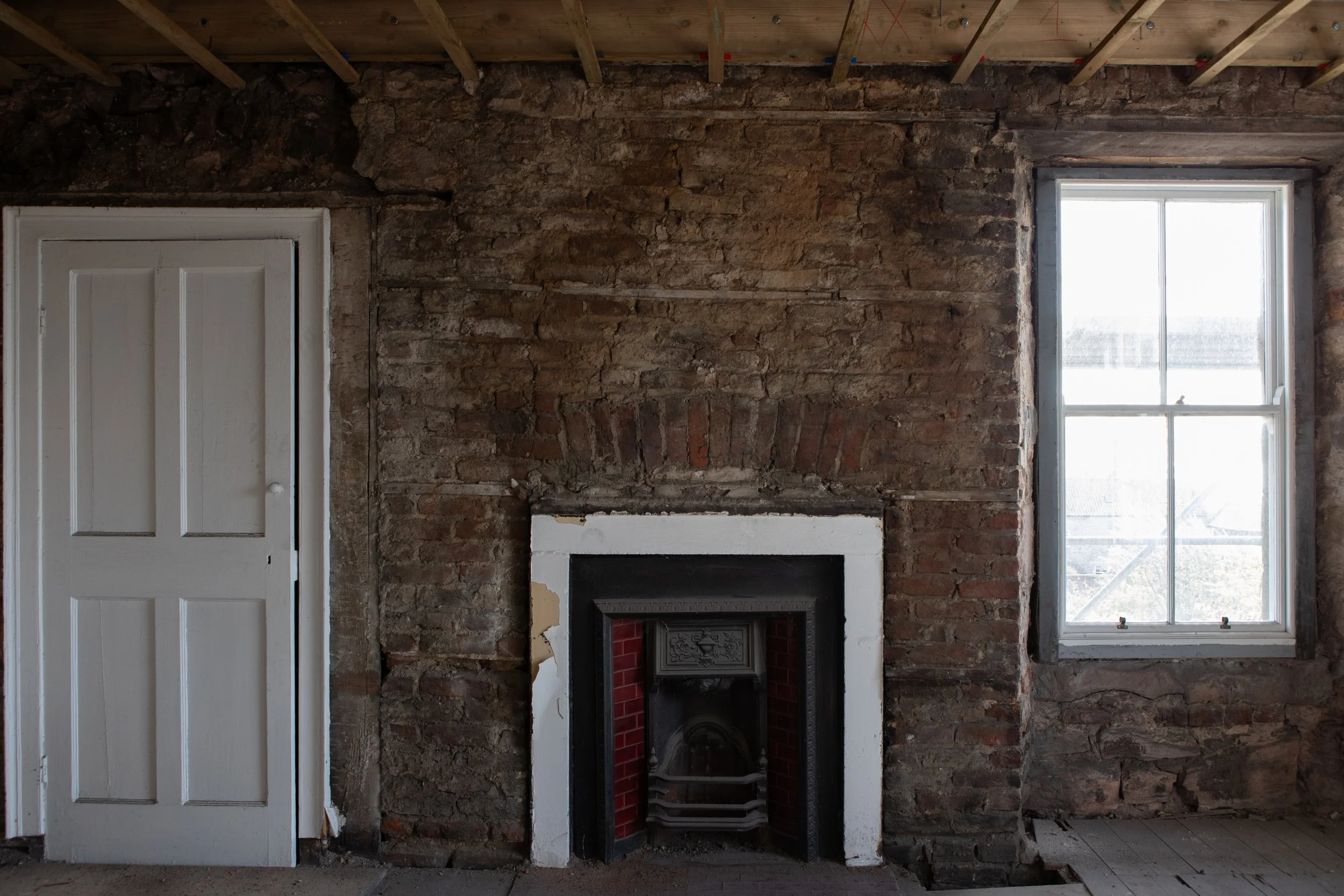 Interior of a room under renovation with partial brick walls, an unfinished ceiling, a white door on the left, and a large window on the right that lets in natural light.