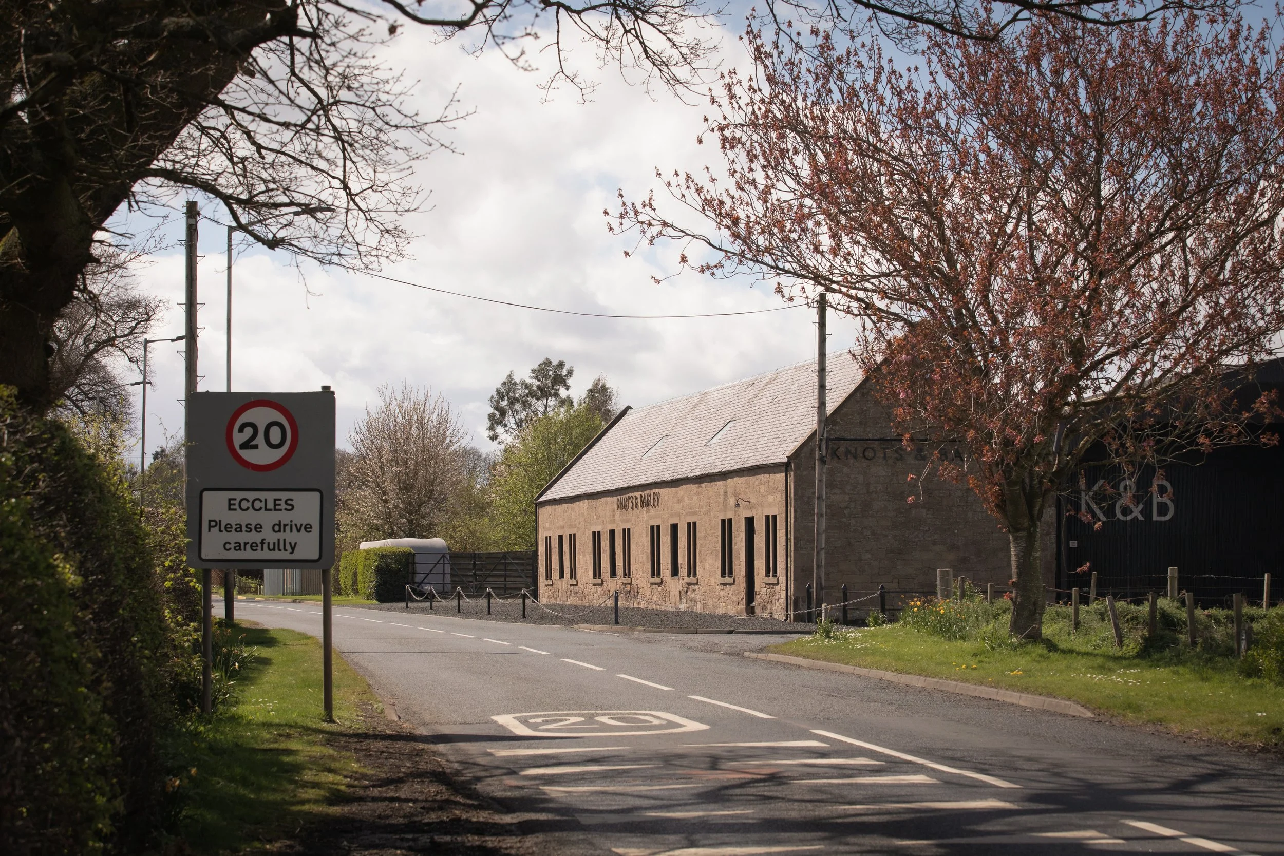 A small street with trees on both sides, some with pink blossoms and others bare. A building with a gray roof and stone walls is visible in the background. There is a speed limit sign indicating 20 mph and a sign for Eagles, instructing drivers to drive carefully.