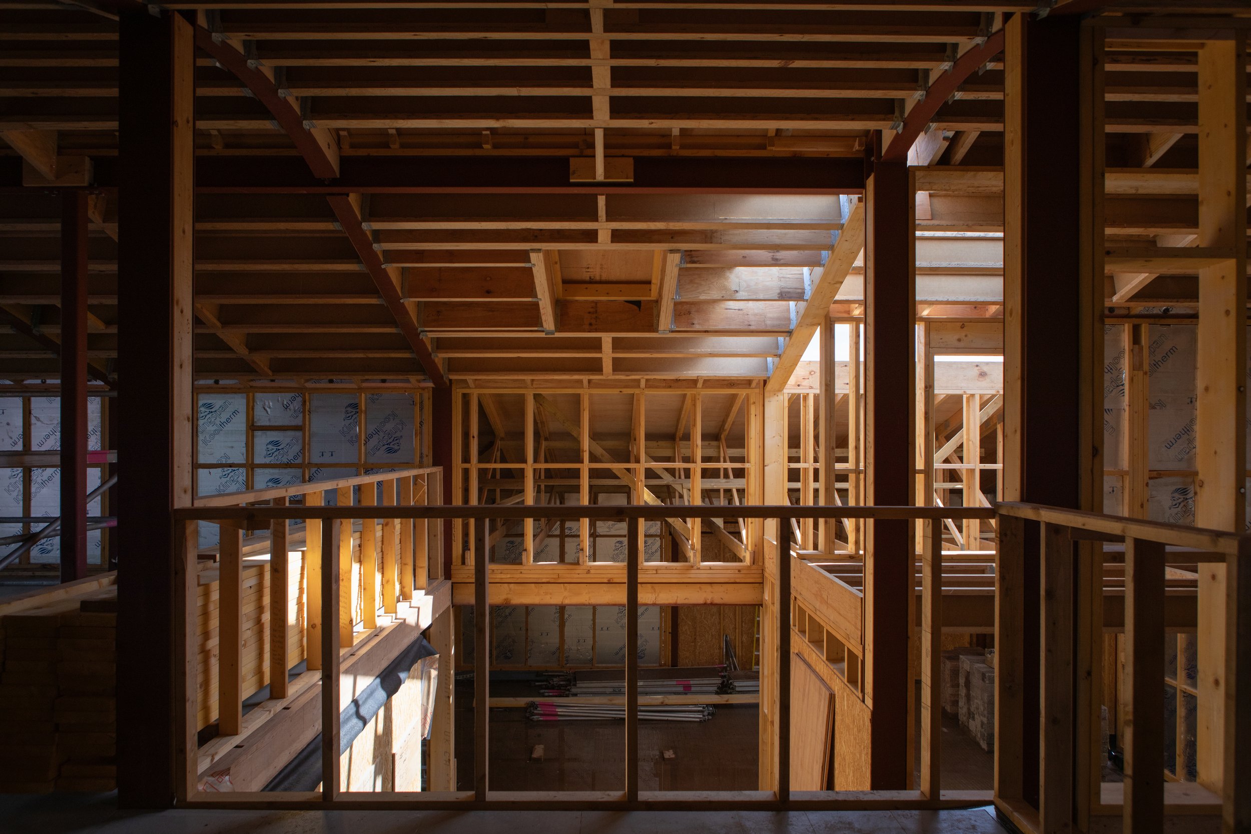 Interior view of a house under construction with exposed wooden framing, beams, and partially built floors and walls.