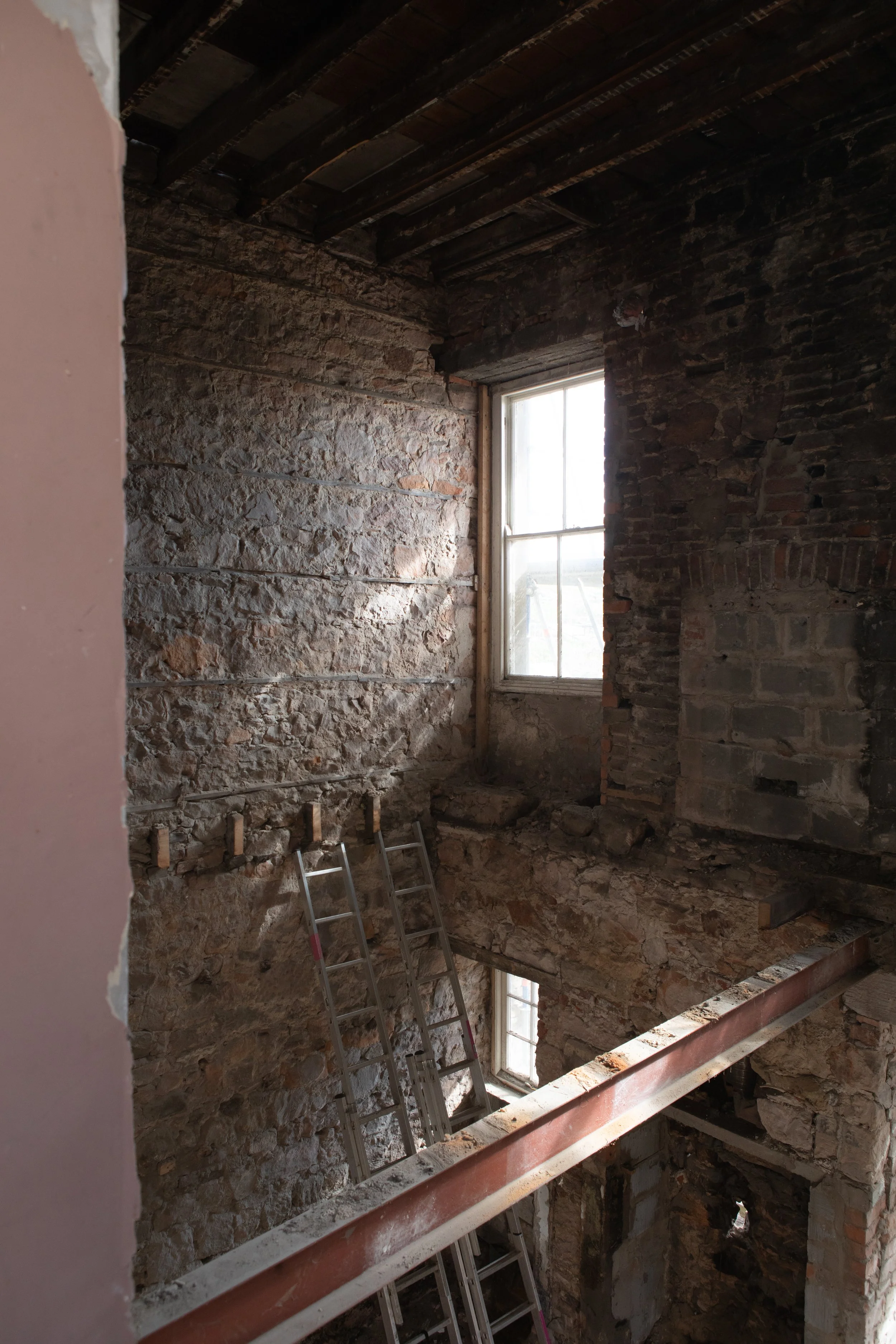 An interior view of a building under renovation with exposed brick and stone walls, a window with sunlight coming through, ladders, and construction beams.
