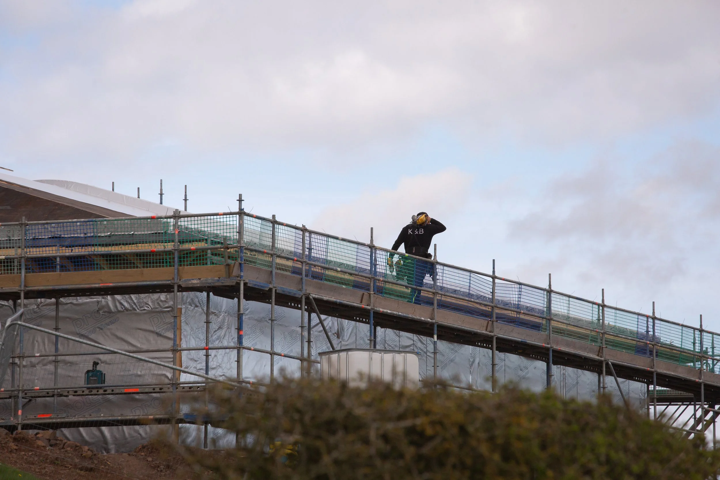 A construction worker stands on a scaffolding at a building site, wearing a black hoodie and a yellow helmet, talking on a cell phone with a cloudy sky overhead.