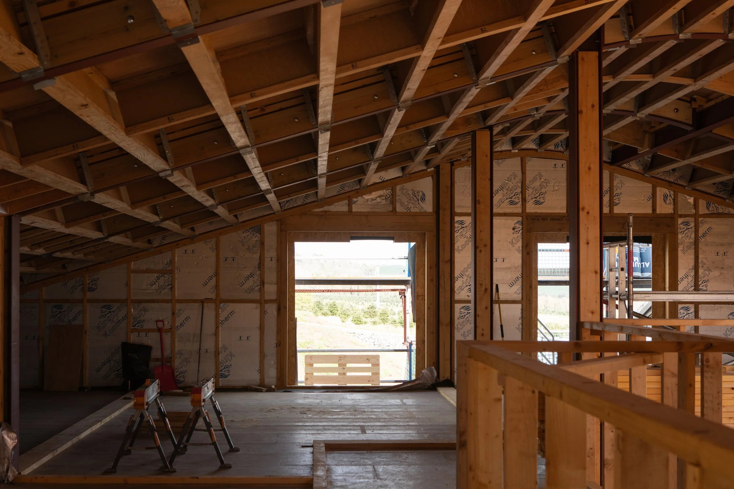 The interior of a house under construction showing wooden framing, with visible roof trusses and window openings, and construction tools on the floor.