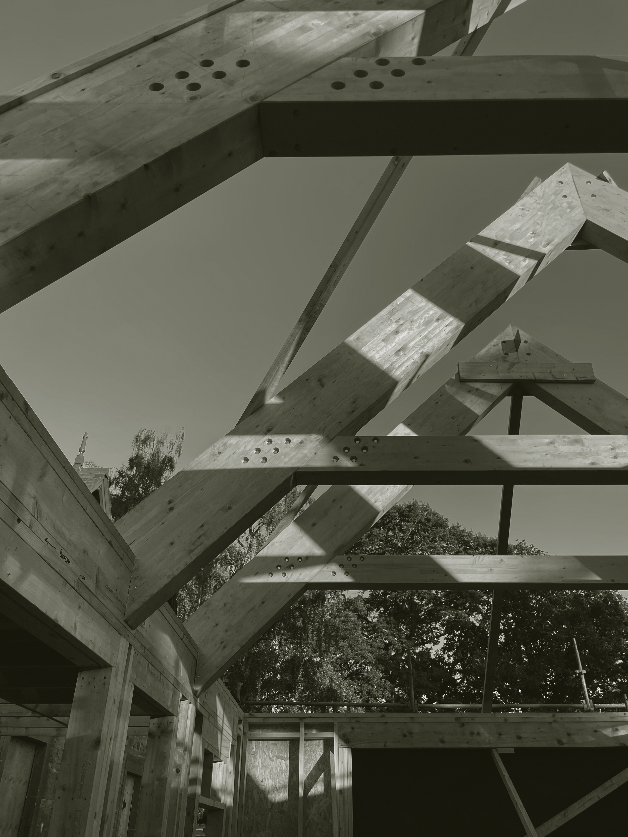 Wooden framing structure of a building under construction, with beams and trusses, and trees in the background.