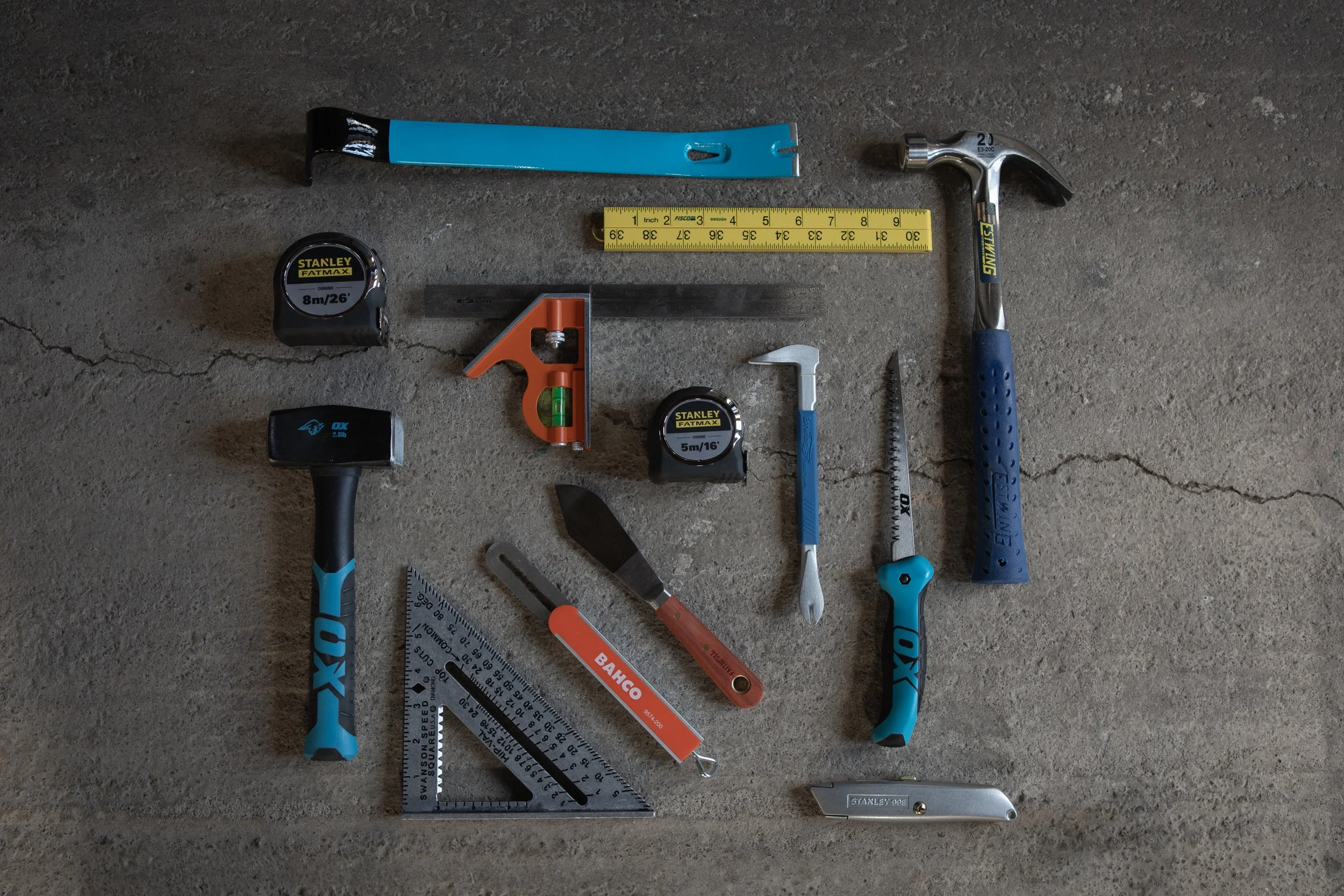 Various measuring and hand tools arranged on a concrete surface, including tape measures, a hammer, a square ruler, a hacksaw, a utility knife, a level, and a putty knife.
