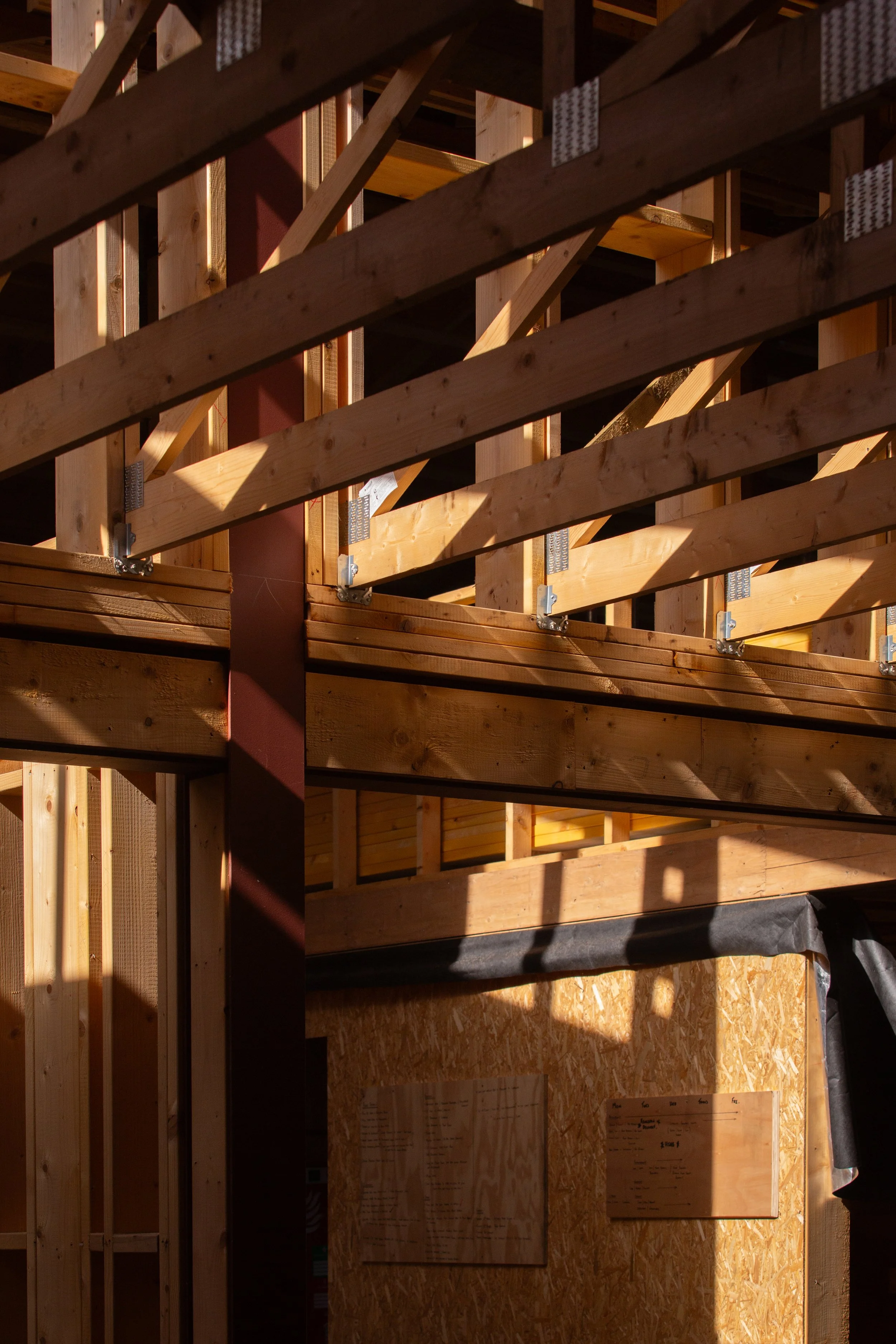 Interior of a building under construction showing wooden framing, vertical studs, diagonal braces, and a plywood wall with plans posted on it.