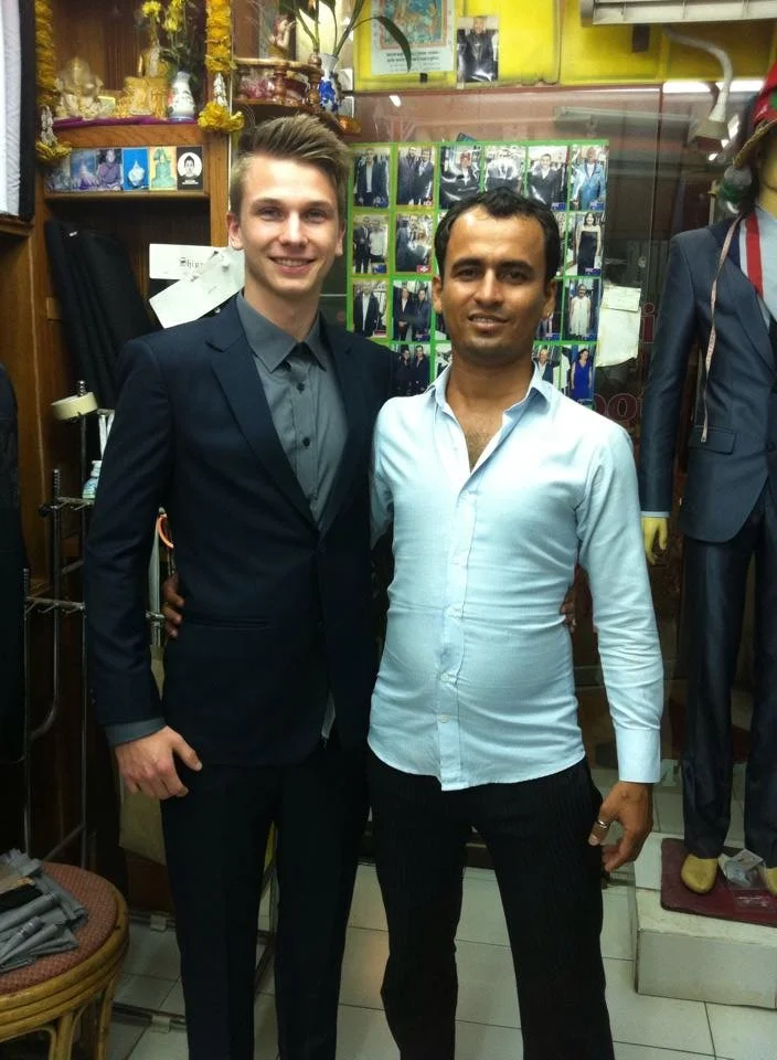 Two men standing side by side, smiling, in a store with mannequins and framed photographs in the background.