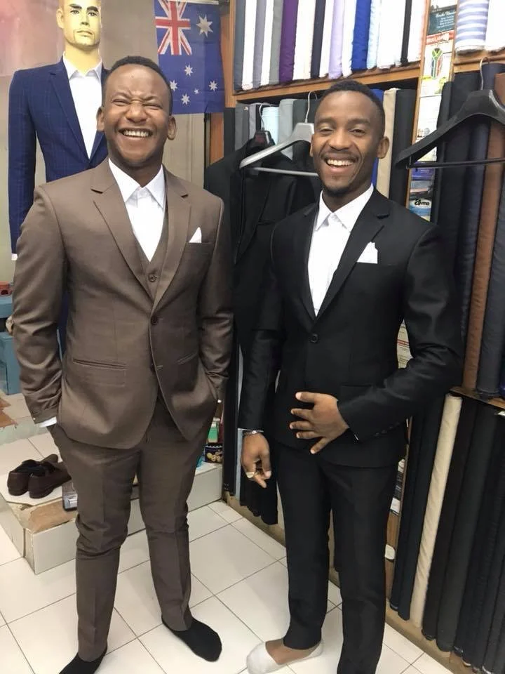 Two men in suits smiling, standing in a fabric store with fabric rolls and an Australian flag in the background.