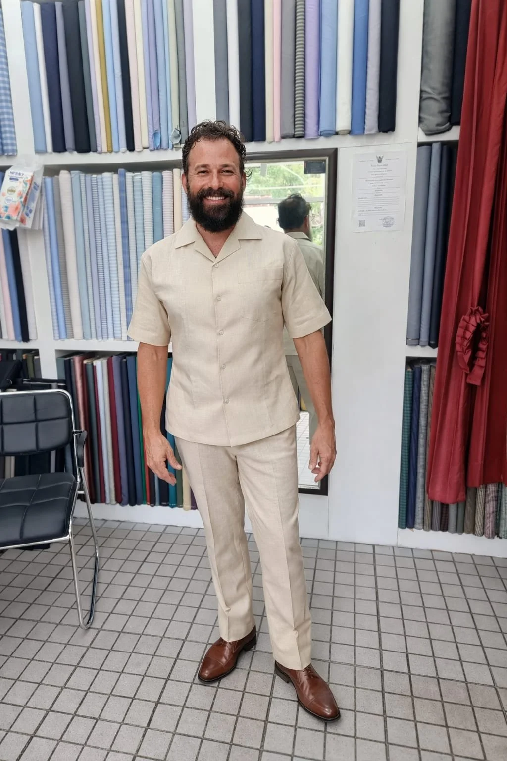 A man with a beard and curly hair smiling, wearing a beige short-sleeved suit, standing in front of shelves filled with fabric bolts in a fabric store.