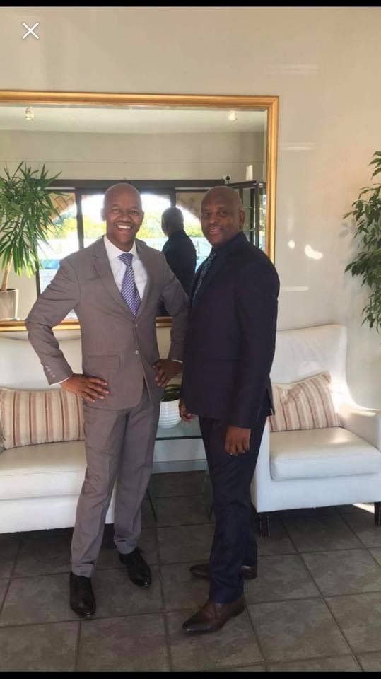 Two men in suits standing in front of a mirror and sofas, smiling at the camera.