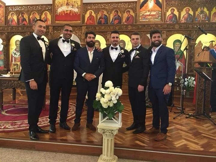 Six men in formal suits standing inside a church with religious icons on the wooden wall behind them, a podium, microphones, and a floral arrangement in the foreground.