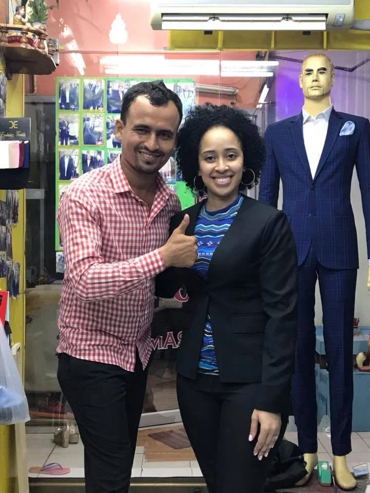 A man and woman smiling and giving a thumbs-up pose in a clothing store, with a mannequin dressed in a navy blue suit in the background.