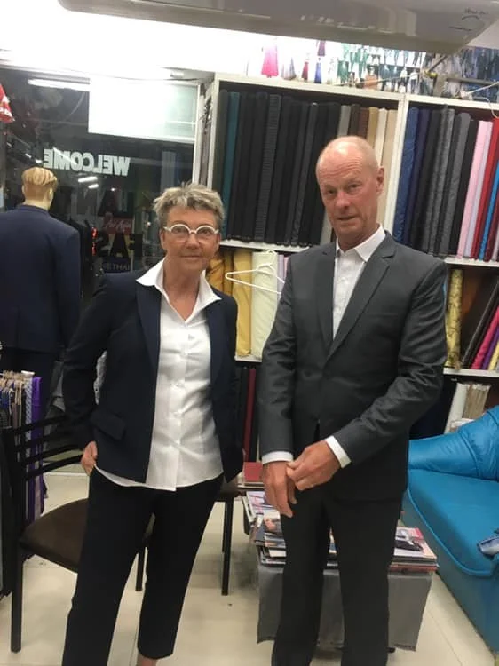 A woman and a man standing in a fabric store or upholstery shop. The woman is wearing glasses, a white blouse, and a dark blazer, while the man is dressed in a gray suit and white shirt. They are posing in front of shelves filled with rolls of fabric