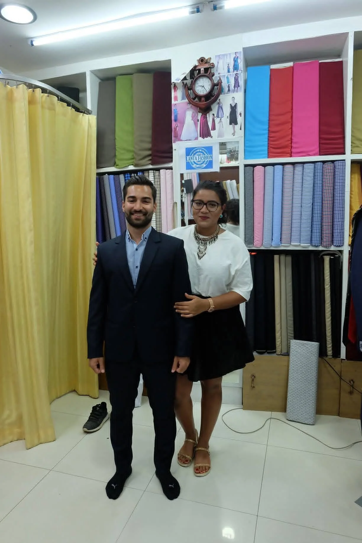 A man and a woman posing together indoors, with fabric rolls on shelves and a yellow curtain in the background.