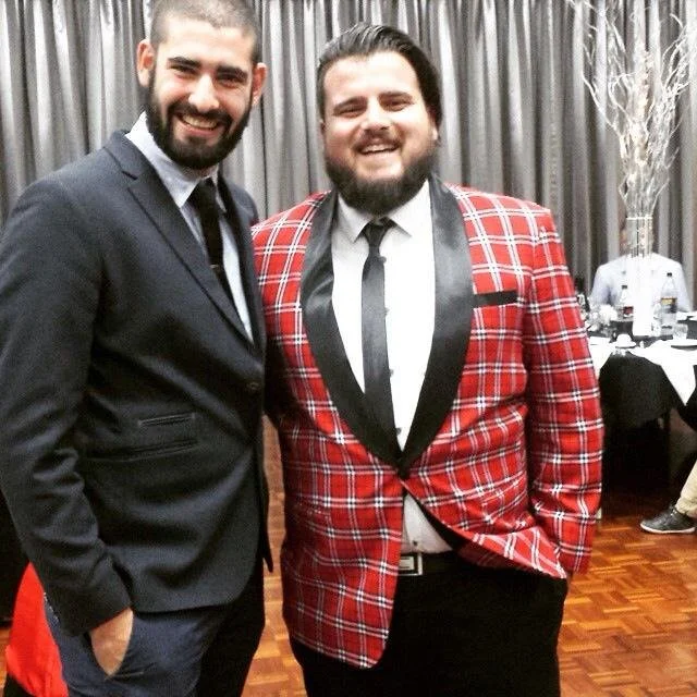 Two smiling men posing together at an indoor event, one in a black suit and the other in a red plaid blazer with black lapels and a black tie.