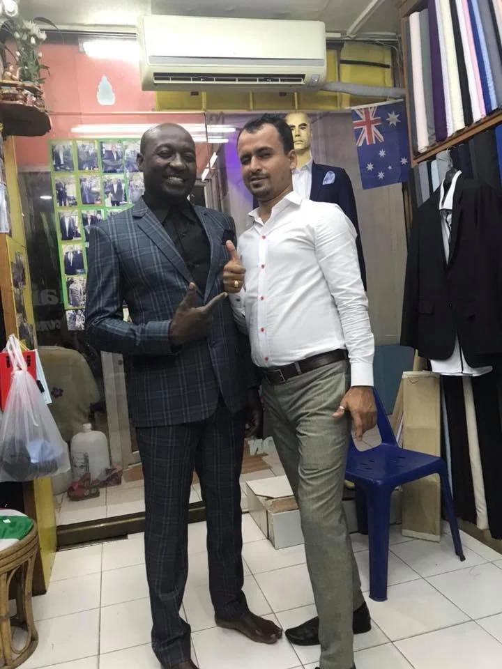 Two men standing in a clothing store, one in a dark plaid suit and photo making a gesture, the other in a white shirt and gray pants, with stitched packaging boxes and clothing racks behind them.