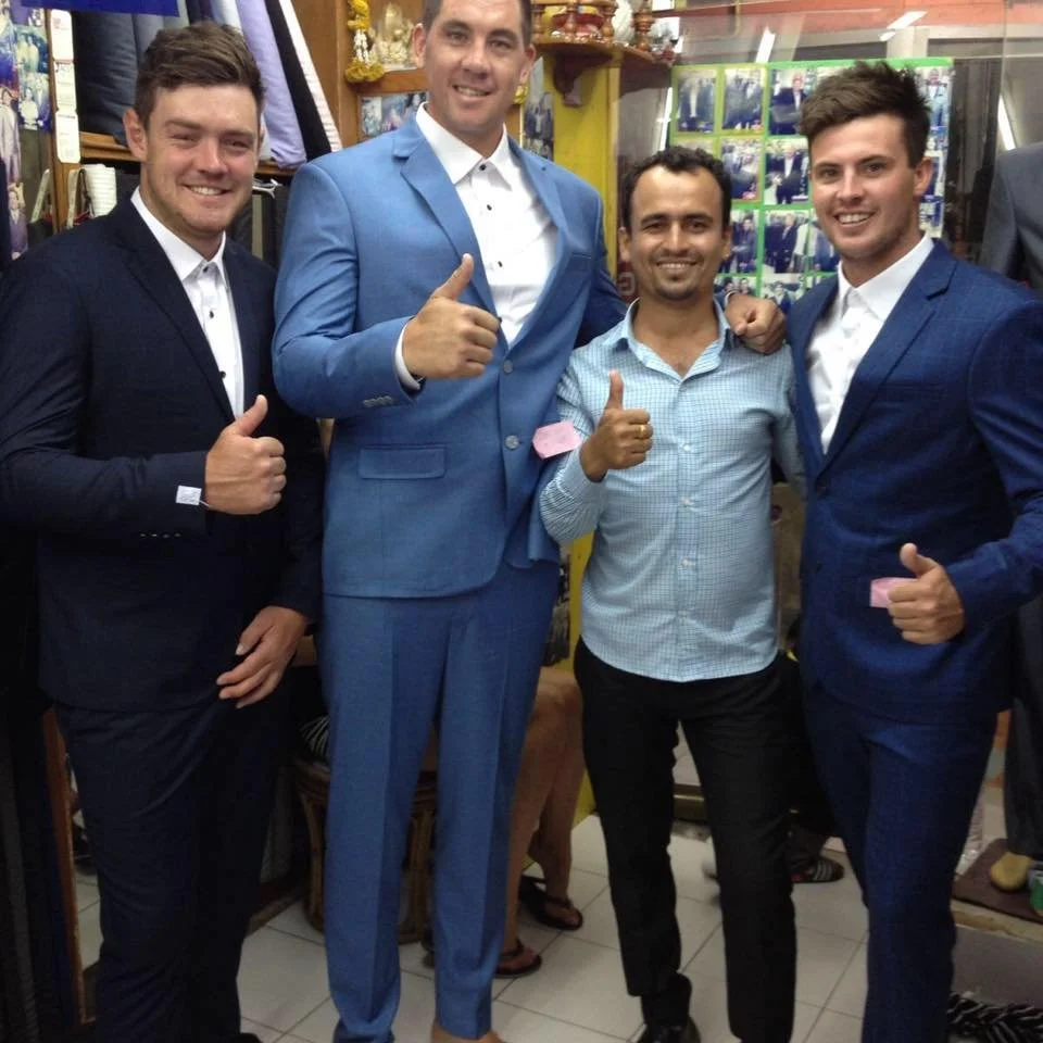 Four men standing together in a store, smiling and giving thumbs up. Two are dressed in suits, one in a light blue suit, and the other in a dark blue suit. The third man is wearing casual clothes, a light blue checkered shirt and black pants. The sto