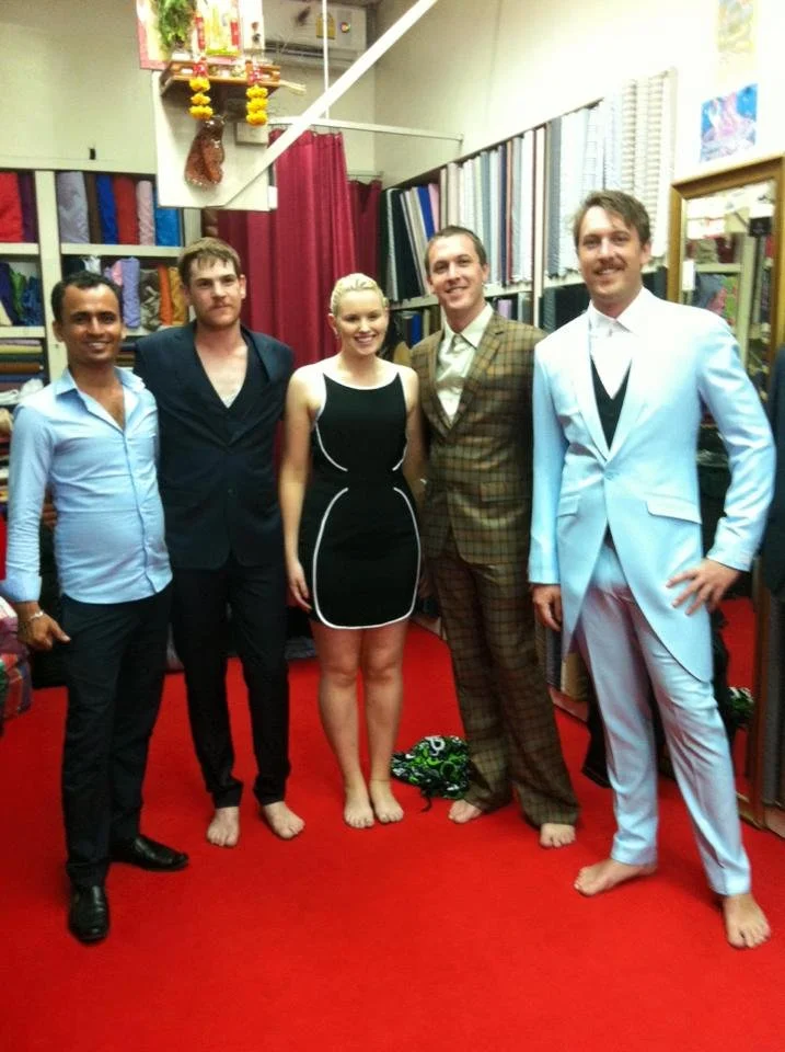 Five people standing barefoot on a red carpet, dressed in formal attire, in a room with shelves of fabric and sewing supplies. They are smiling and posing for the photo.