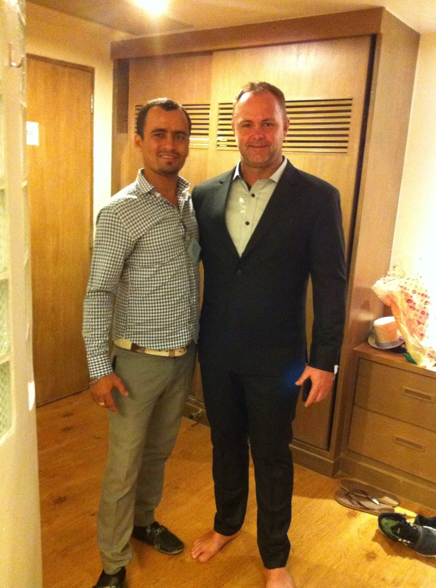 Two men standing together indoors, one in casual attire and the other in a black suit and white shirt, smiling at the camera.
