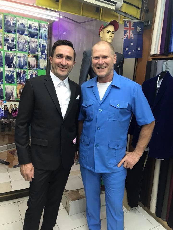 Two men standing side by side in a store. One is wearing a black suit, white shirt, and smiling; the other is in a blue uniform with a white shirt, also smiling. Behind them, there is a mannequin, an Australian flag, and a display of photos.