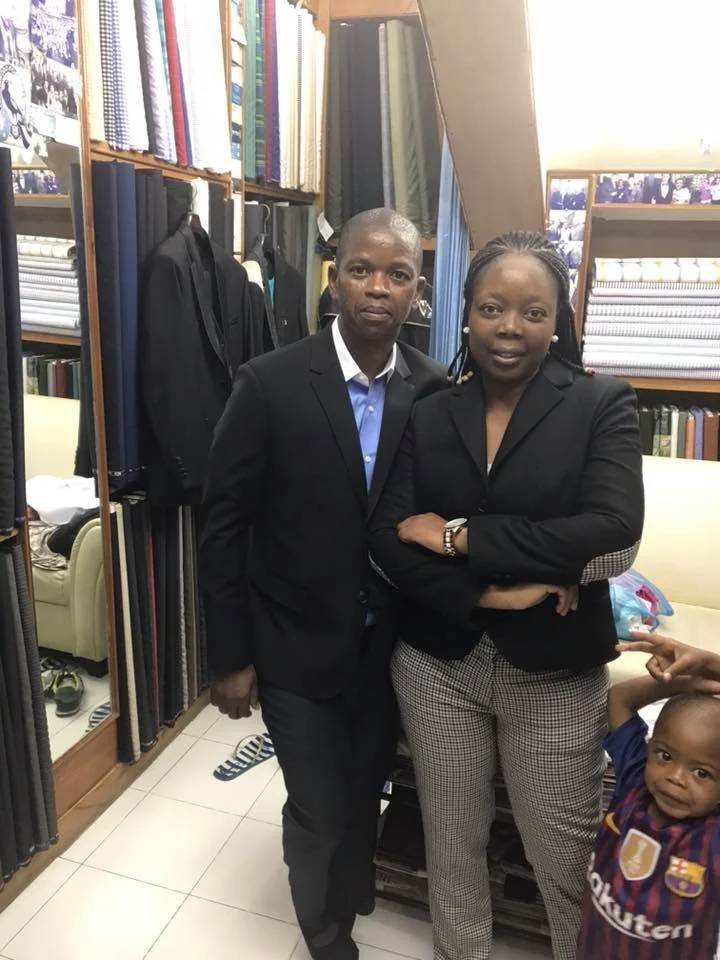 Two adults and one child standing indoors in a clothing store or tailor shop, with shelves of fabric and garments behind them. The adults are dressed in black blazers, and the child is wearing a sports jersey.