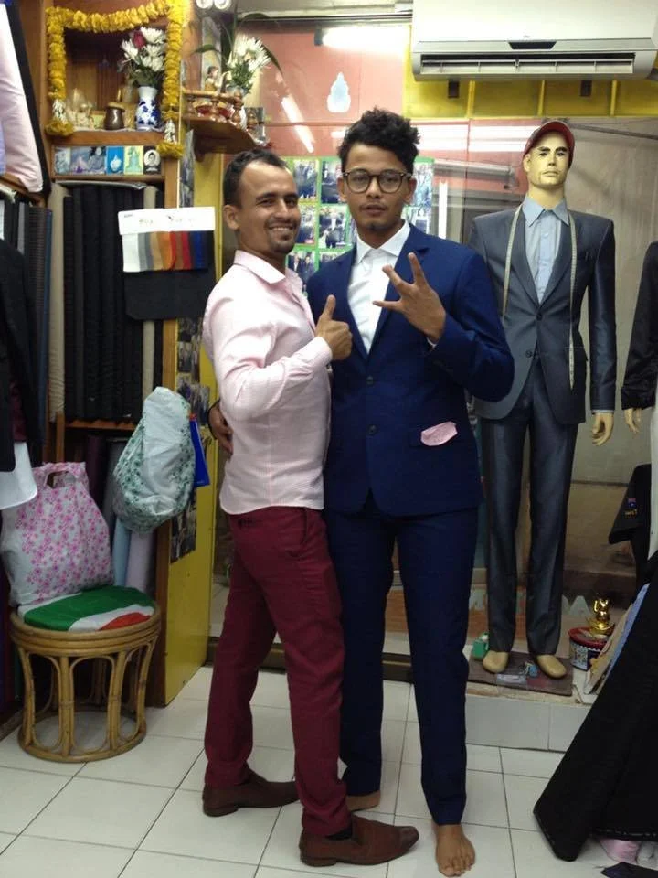 Two men standing together inside a clothing store, one in a pink shirt and red pants, the other in a blue suit and glasses, posing for the photo. A mannequin in a suit and tie is visible in the background.