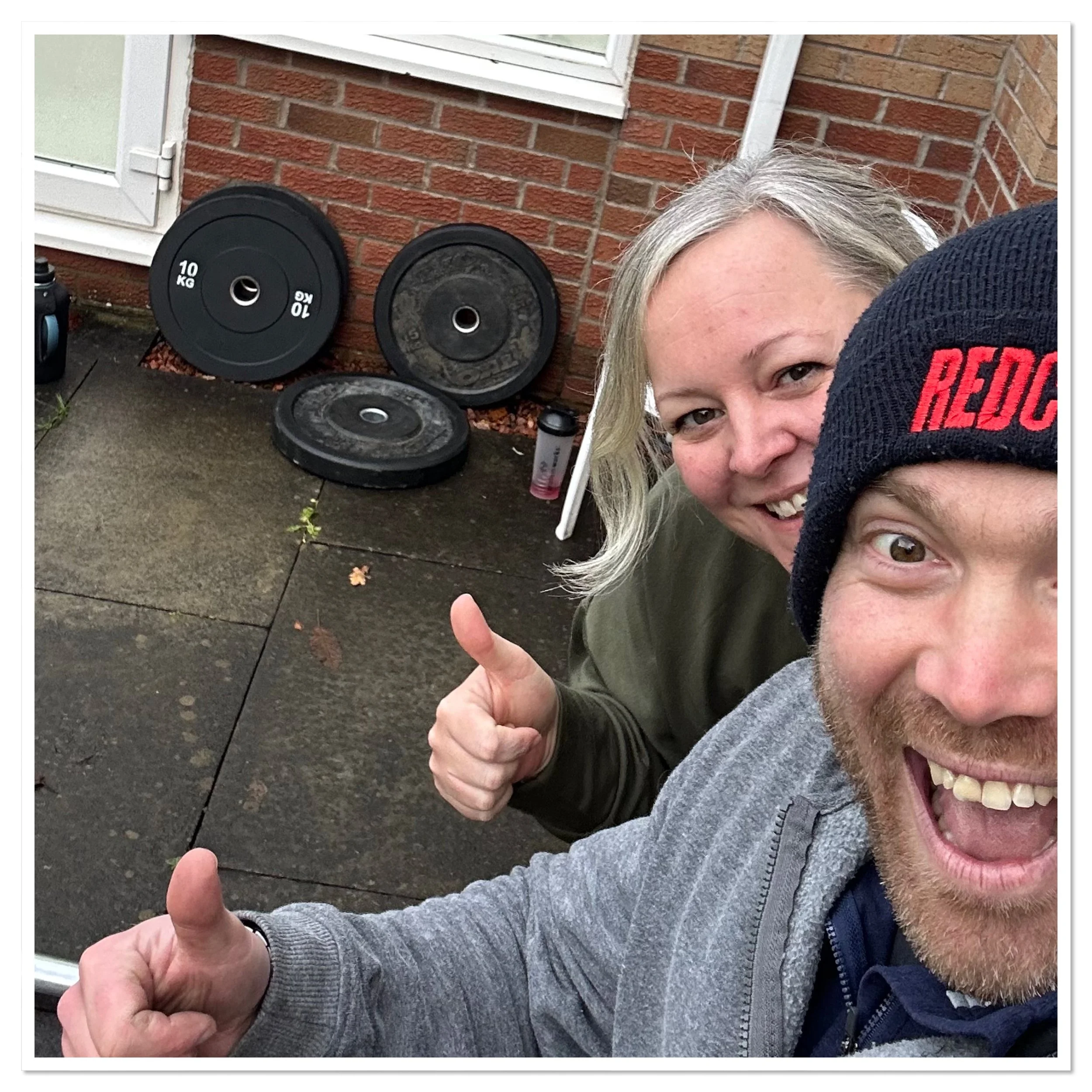 Smiling man and woman taking a selfie outdoorsnafter a personal training session with gym weights and a water bottle in the background.