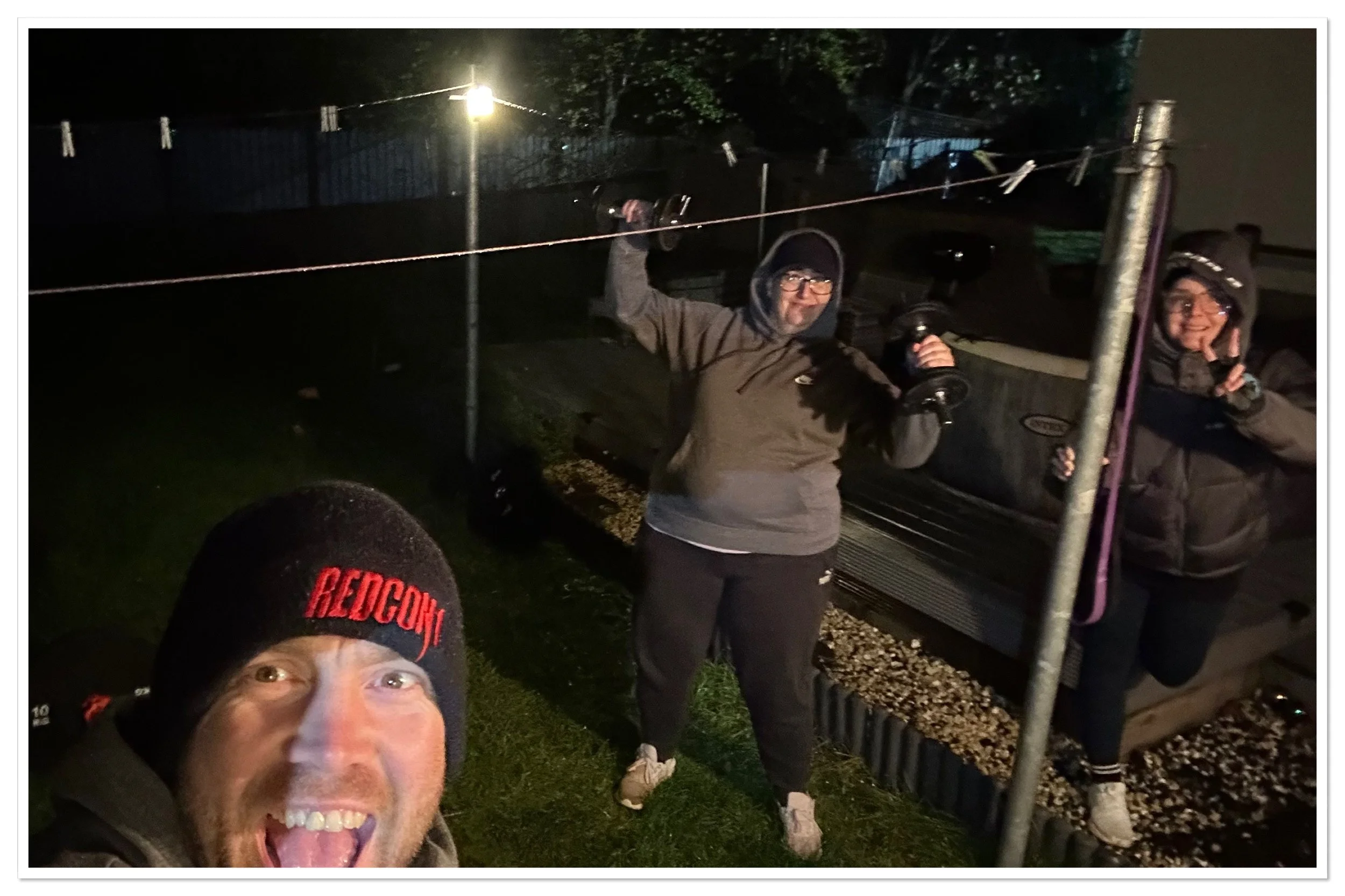 Outdoors personal training session at night, with two holding dumbbells and one taking a selfie, dressed in warm clothing including hats and hoodies