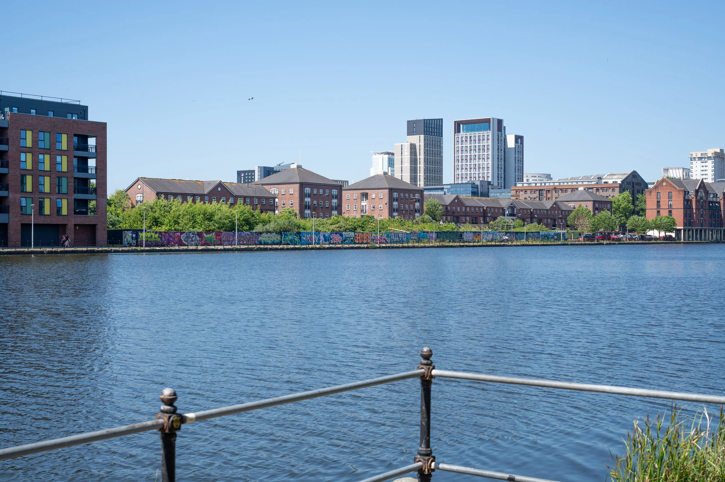 Cityscape across a river with modern and historic buildings, blue sky, and graffiti on a wall along the riverbank.