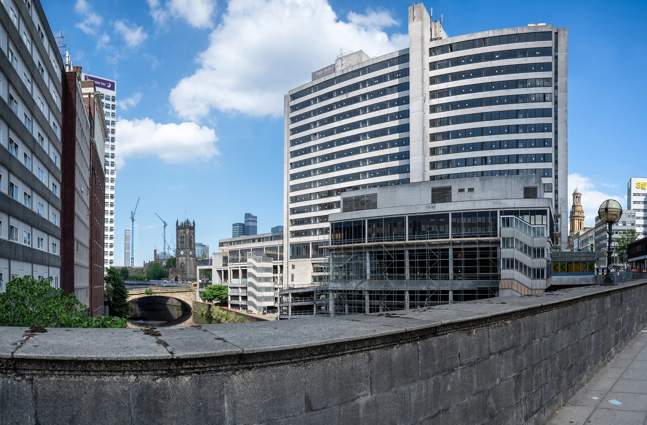 Cityscape with tall modern buildings, a stone wall in the foreground, and a partly cloudy blue sky.