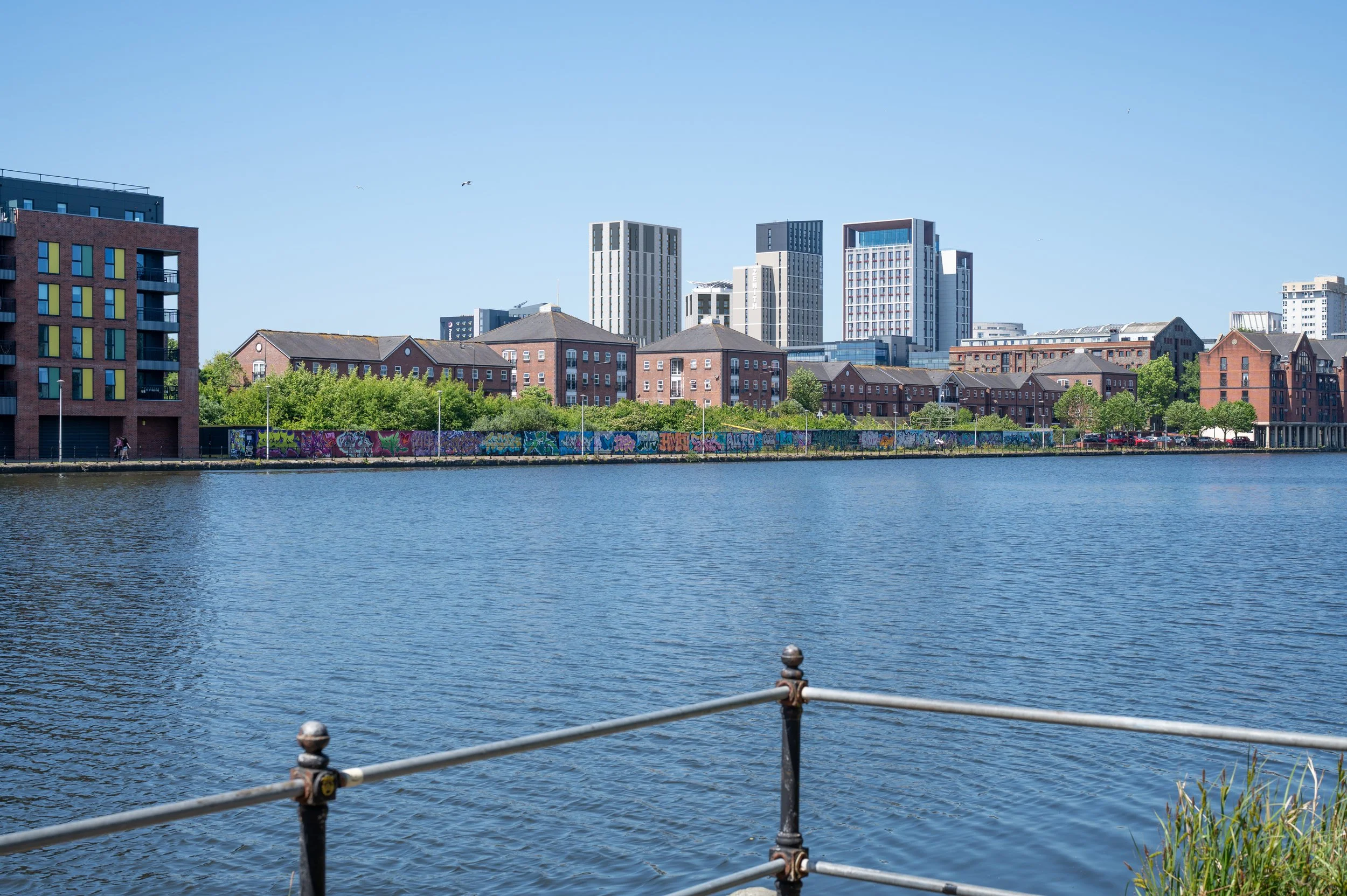 City skyline with modern buildings and older brick structures, waterfront with railing, and graffiti on a fence, under clear blue sky.