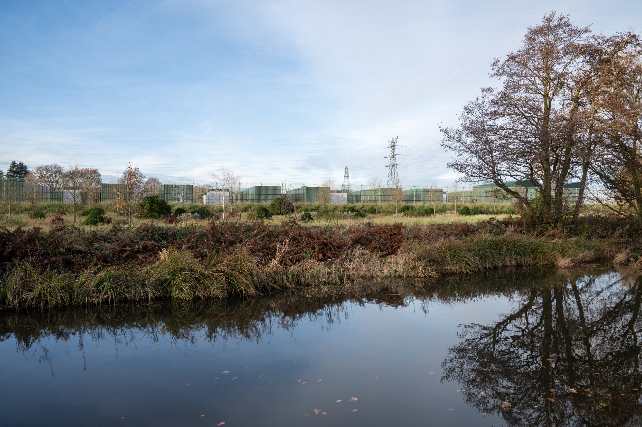 A landscape with a small body of water in the foreground, grassy and shrubby terrain behind it, and industrial buildings with power lines in the background under a partly cloudy sky.